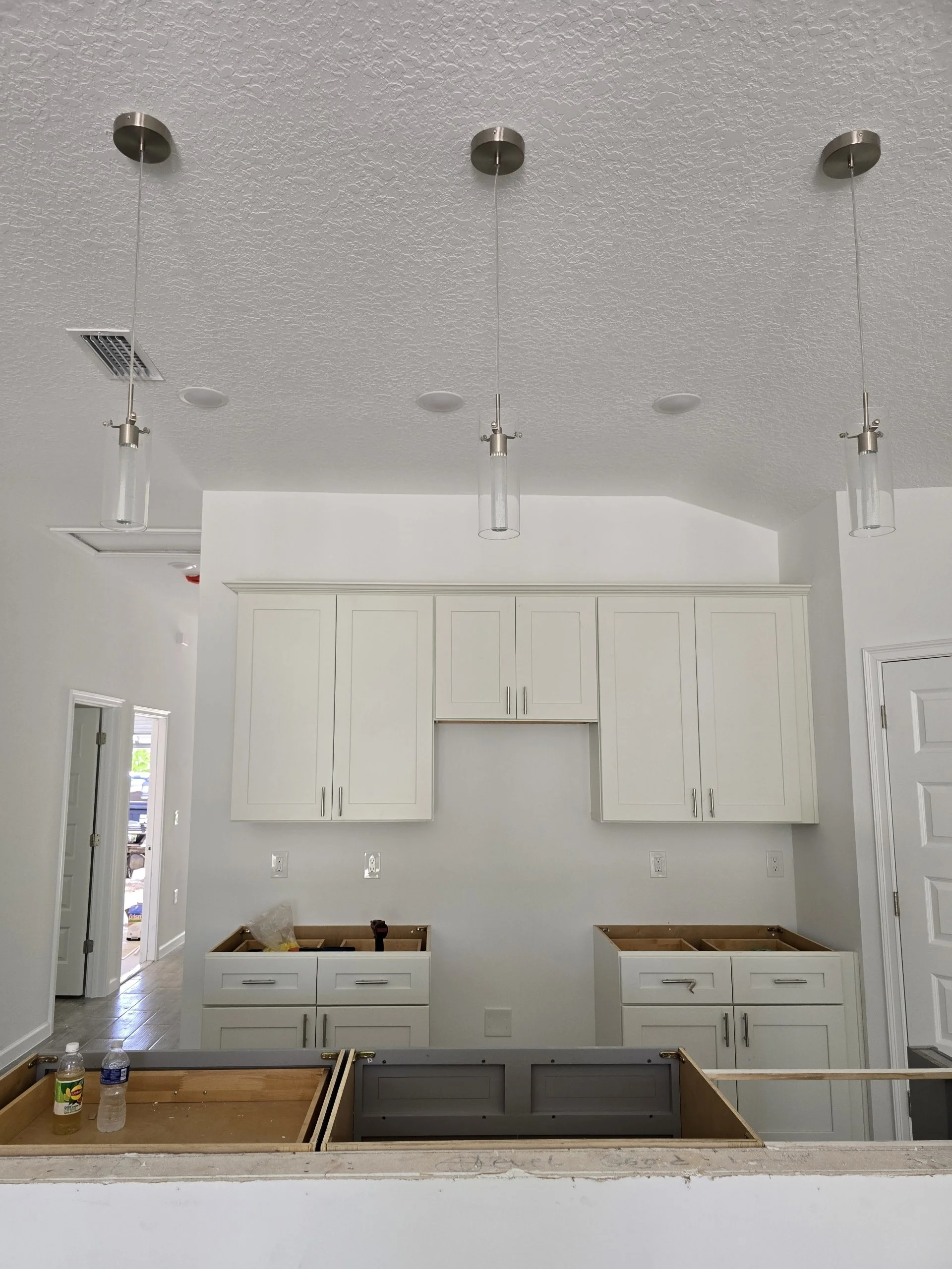 Kitchen under construction with white cabinets, ceiling lights, and an unfinished countertop with water bottles and a lemon soda.