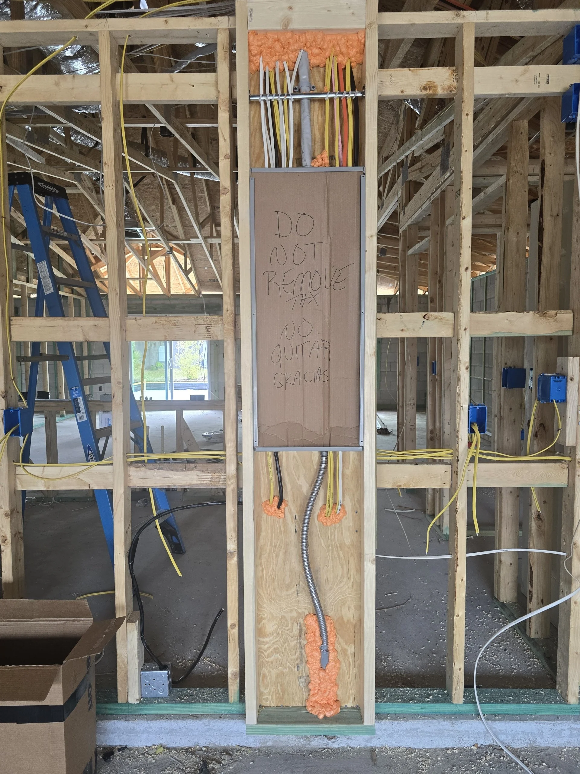 A construction site with exposed wooden framing, electrical wiring, and a handwritten sign that says "DO NOT REMOVE THIS NO QUITAR" on a piece of cardboard.