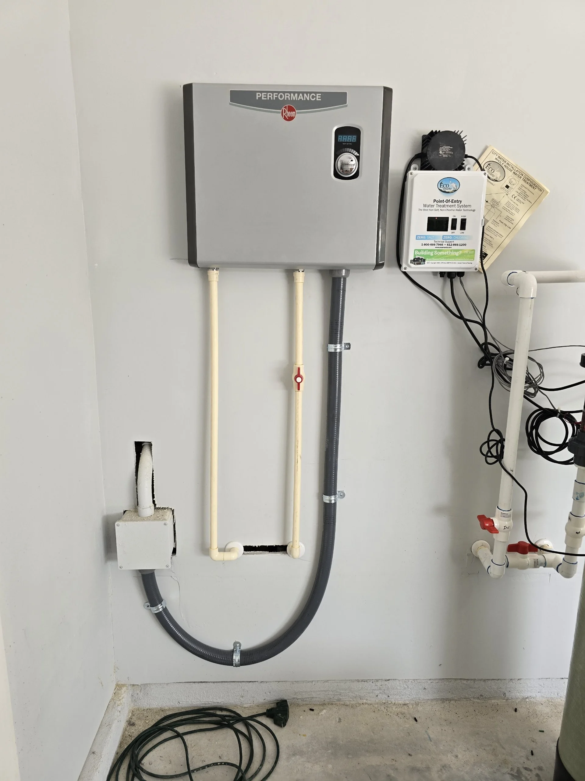 Water treatment system with gray control box, pipes, and electronic components on a white wall.