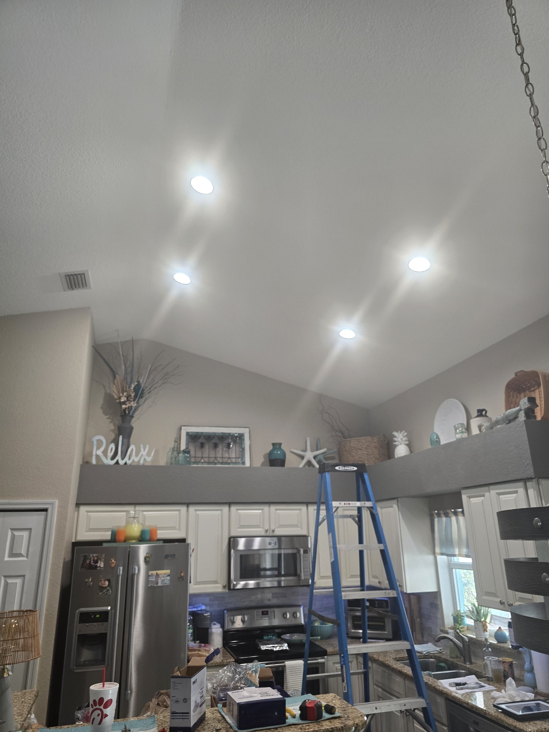 Kitchen with ceiling lights, shelves with decorative items, a ladder, refrigerator, microwave, and countertop clutter.