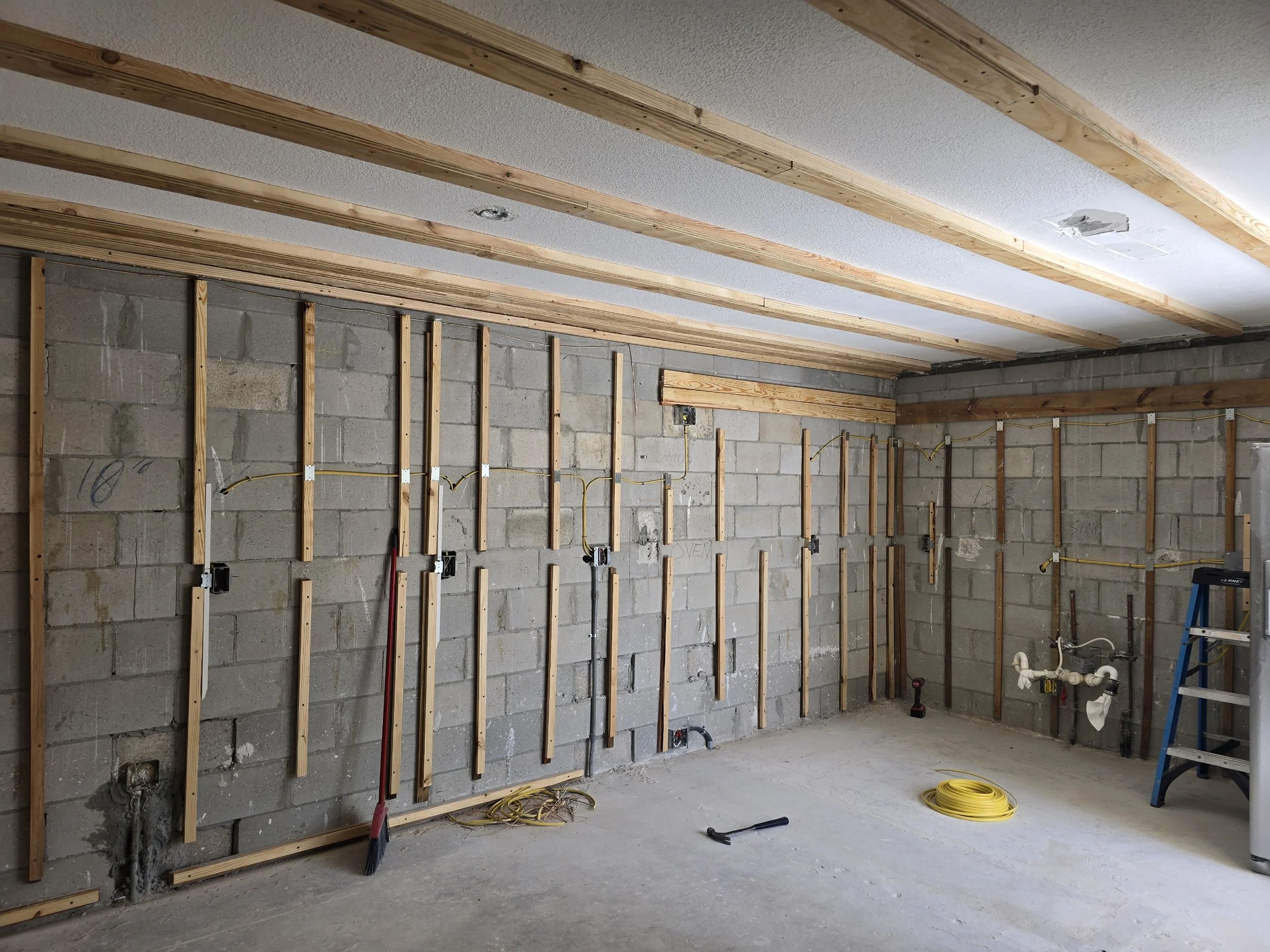 Interior of a room under construction with exposed wooden studs on a concrete block wall and electrical wiring, a ladder, and construction tools.