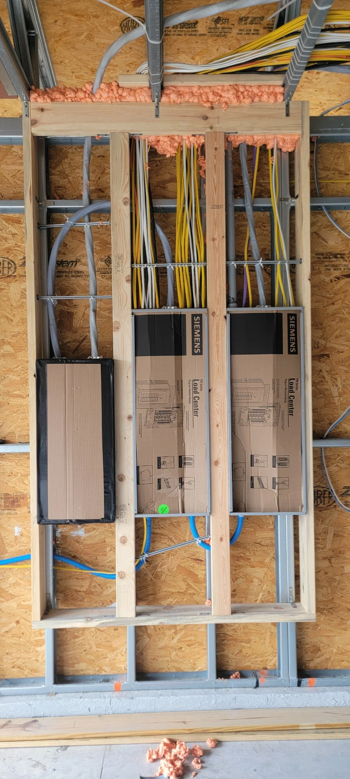 Electrical wiring installation in progress within a wooden framing structure. Includes conduits, cables, load center boxes, and insulation foam at the top.