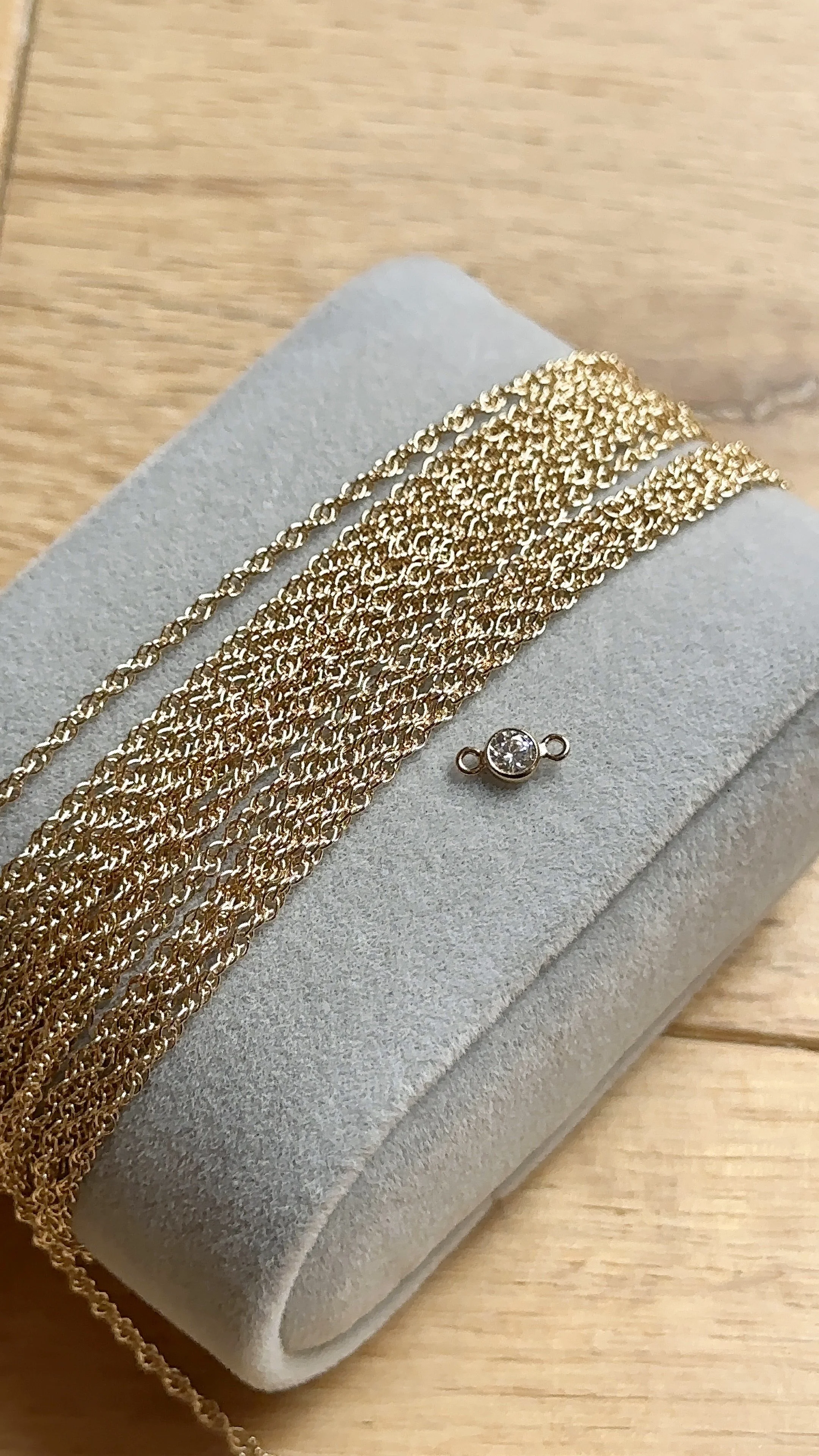 Macro photo of a collection of delicate gold rope chains fanned out on a grey velvet pad, shown next to a small, bezel-set cubic zirconia charm with two loops for permanent jewellery linking.