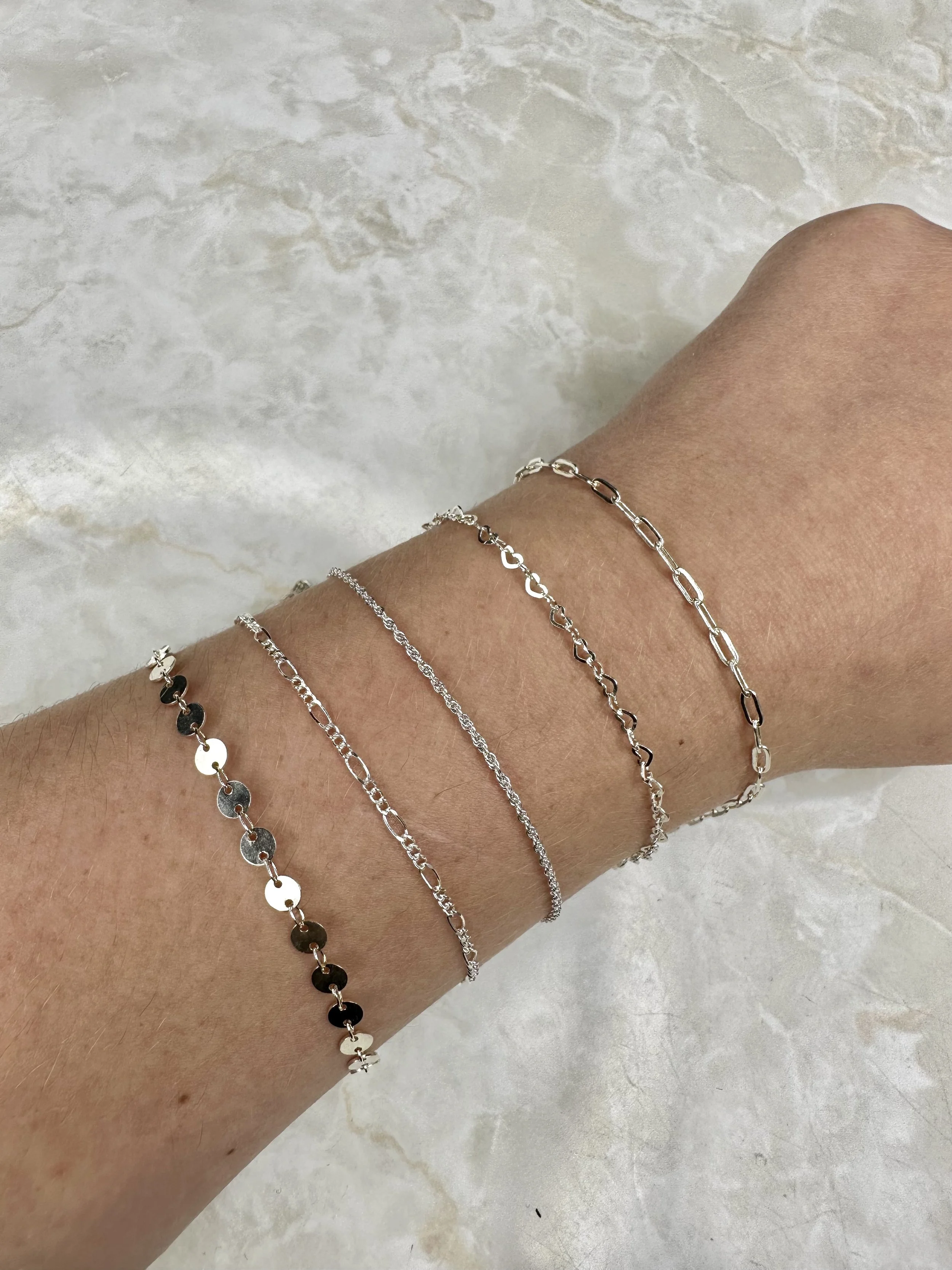 A collection of five 925 sterling silver permanent bracelets on a wrist, featuring various delicate chain styles including paperclip, heart-link, rope, figaro, and flat sequin (disc) chains.