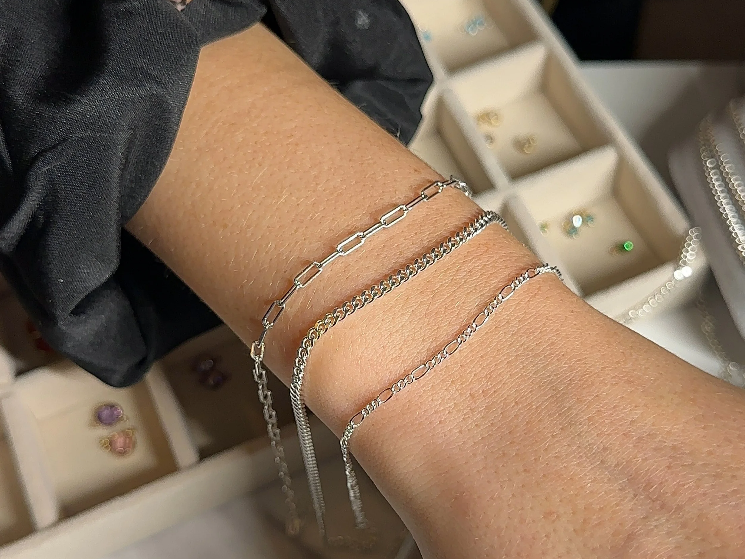 Close-up of three permanent sterling silver bracelets on a wrist, featuring paperclip, curb, and delicate figaro links, with a jewellery tray visible in the background.