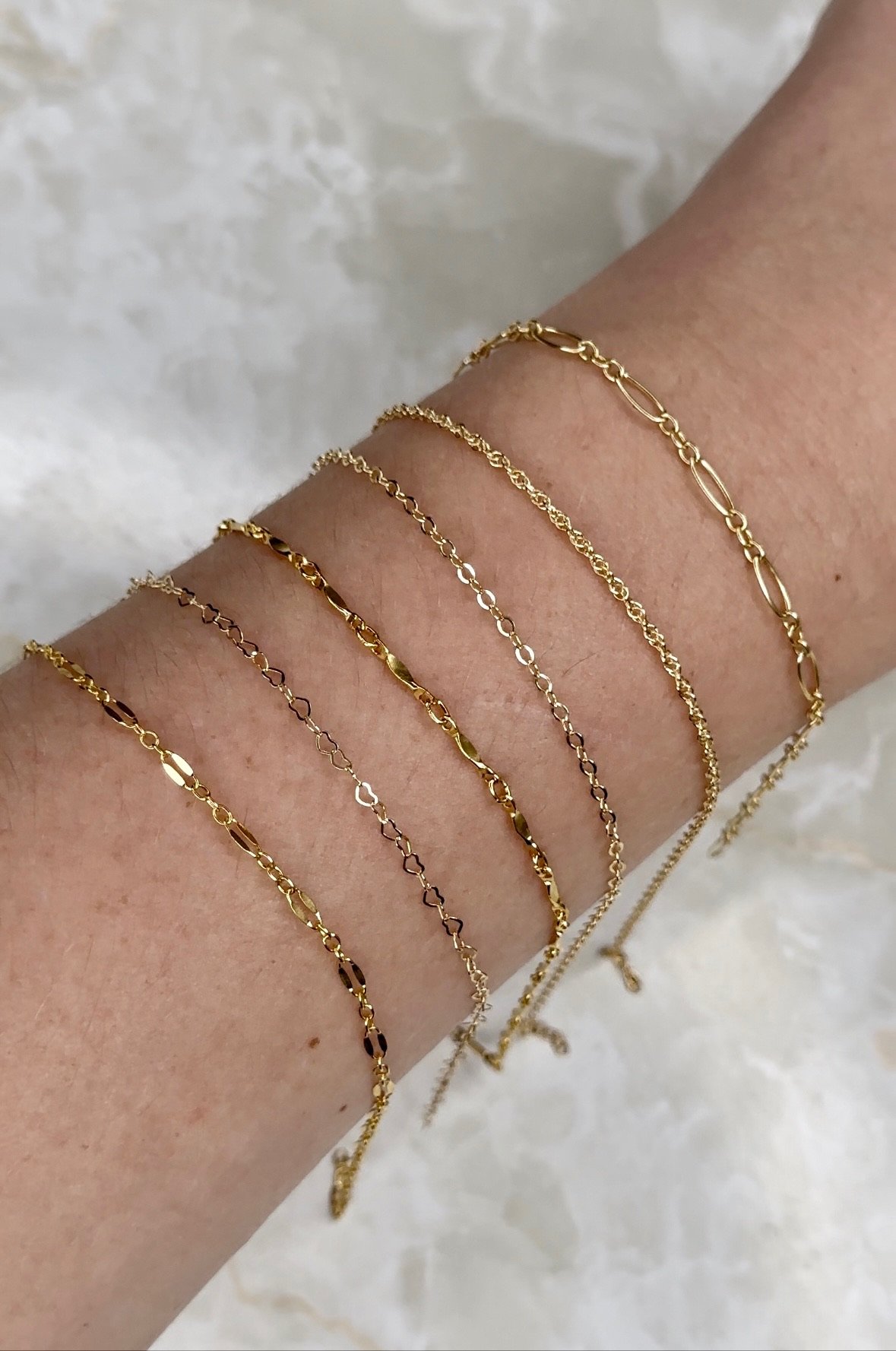 Close-up of six delicate gold chains, including figaro, heart-link, and twist designs, being worn as permanent bracelets on a person's wrist.