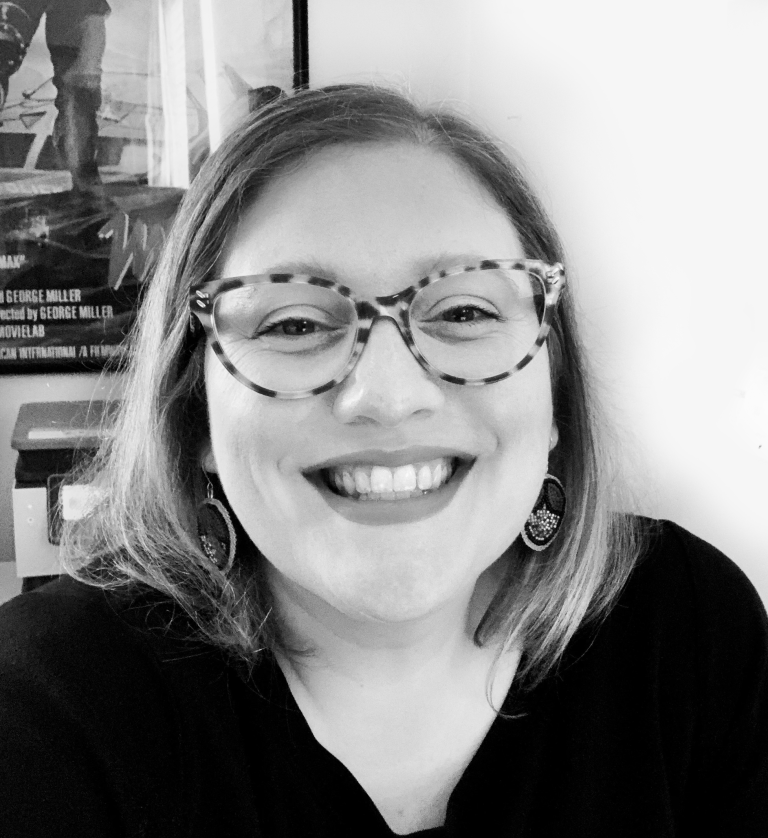 A smiling woman with shoulder-length hair, wearing patterned glasses and earrings, in a black top, indoors with a poster in the background.