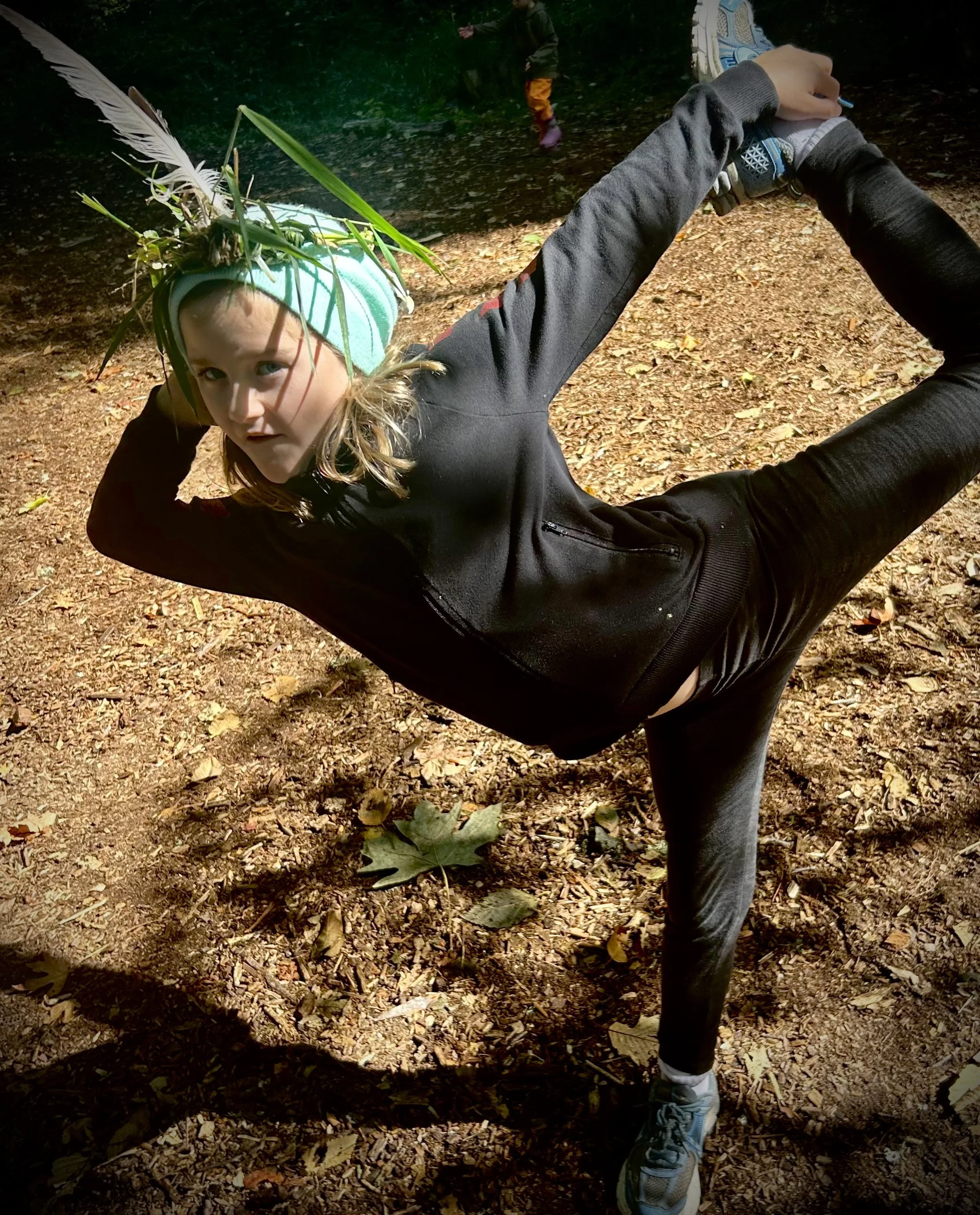 A girl in black athletic clothing balancing on one leg with her other leg raised and holding her foot, outdoors on a dirt trail with fallen leaves, with a person in the background wearing orange pants and purple shoes.