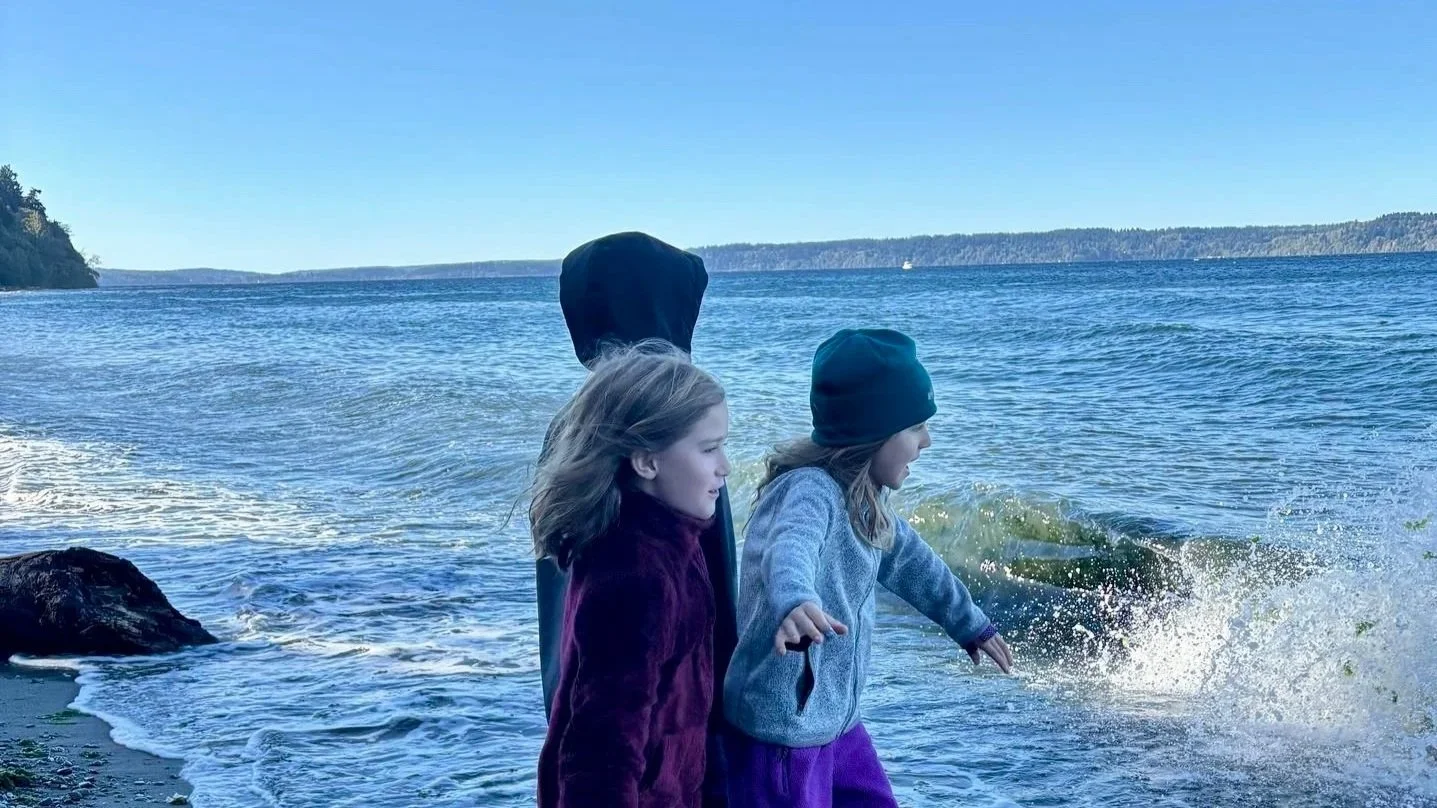 Two young girls wearing jackets and beanie hats playing by the water, with one girl splashing in the ocean.