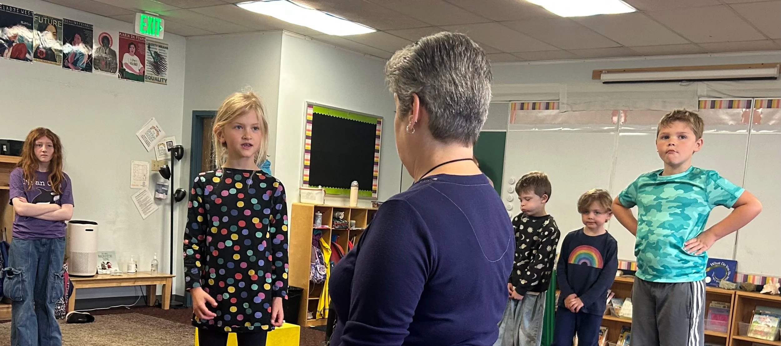 A classroom with children standing in a line, a teacher facing them, and colorful posters on the wall.
