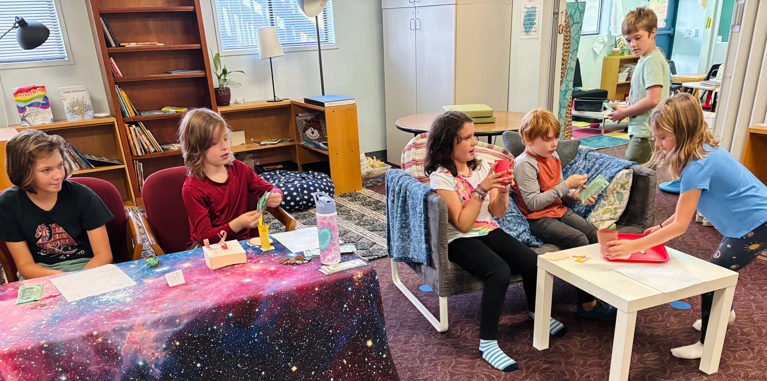Children in a classroom participating in an activity with some seated at a table and others standing and handing out cards or papers.