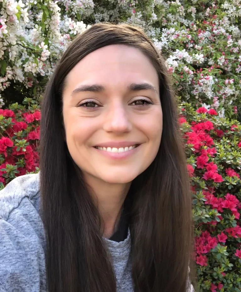 A young woman with long brown hair smiling in front of blooming pink, white, and purple flowers.