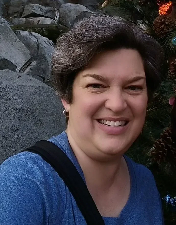 A woman with short dark hair smiling outdoors, wearing a blue shirt and a black strap over her shoulder with pine cones and Christmas lights in the background.