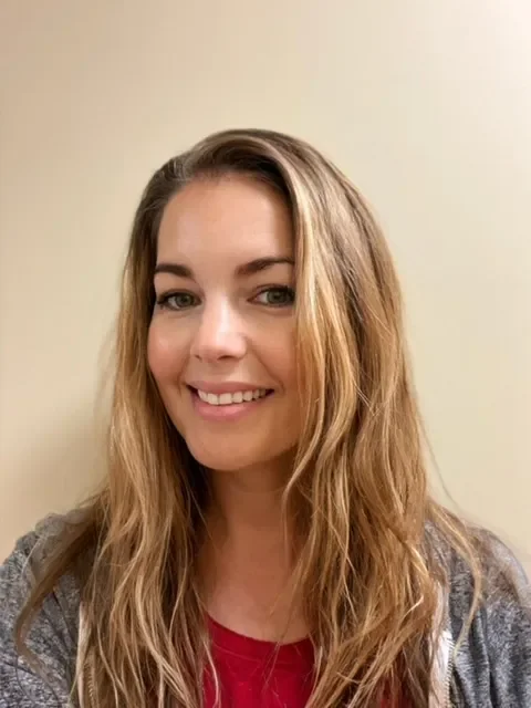 Portrait of a young woman with long, wavy blonde hair, smiling, wearing a red top and gray hoodie, standing against a beige wall.