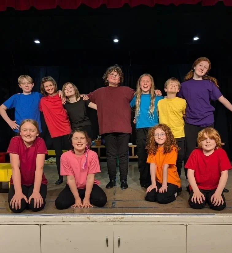 Group of children on stage, posing for a photo, wearing colorful t-shirts in red, blue, pink, yellow, orange, and purple.