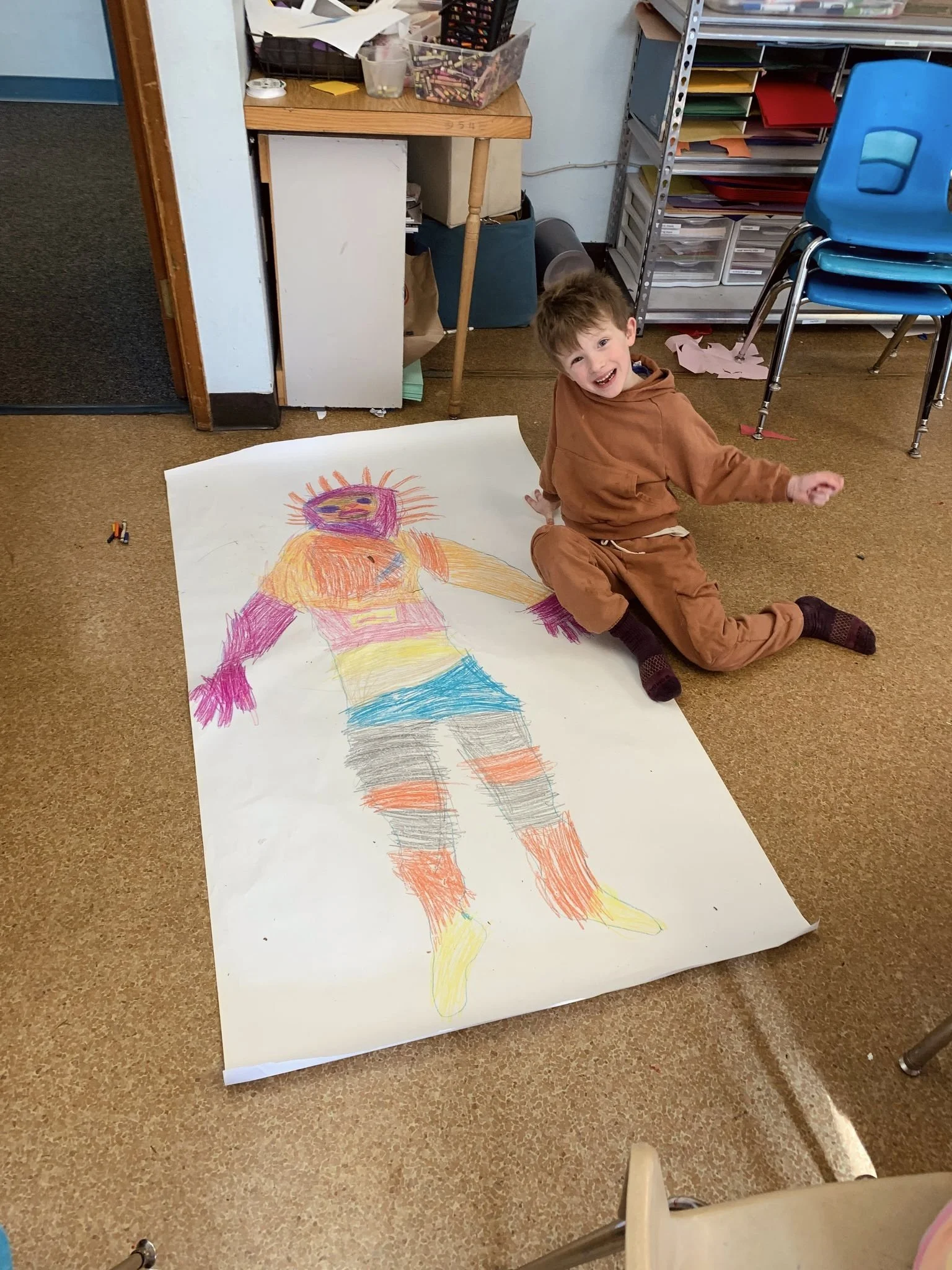 A young boy in a brown hoodie and brown pants sitting on the floor next to a large colorful crayon drawing of a person on a big sheet of paper. The boy is smiling and looking at the camera in a classroom or art room setting.