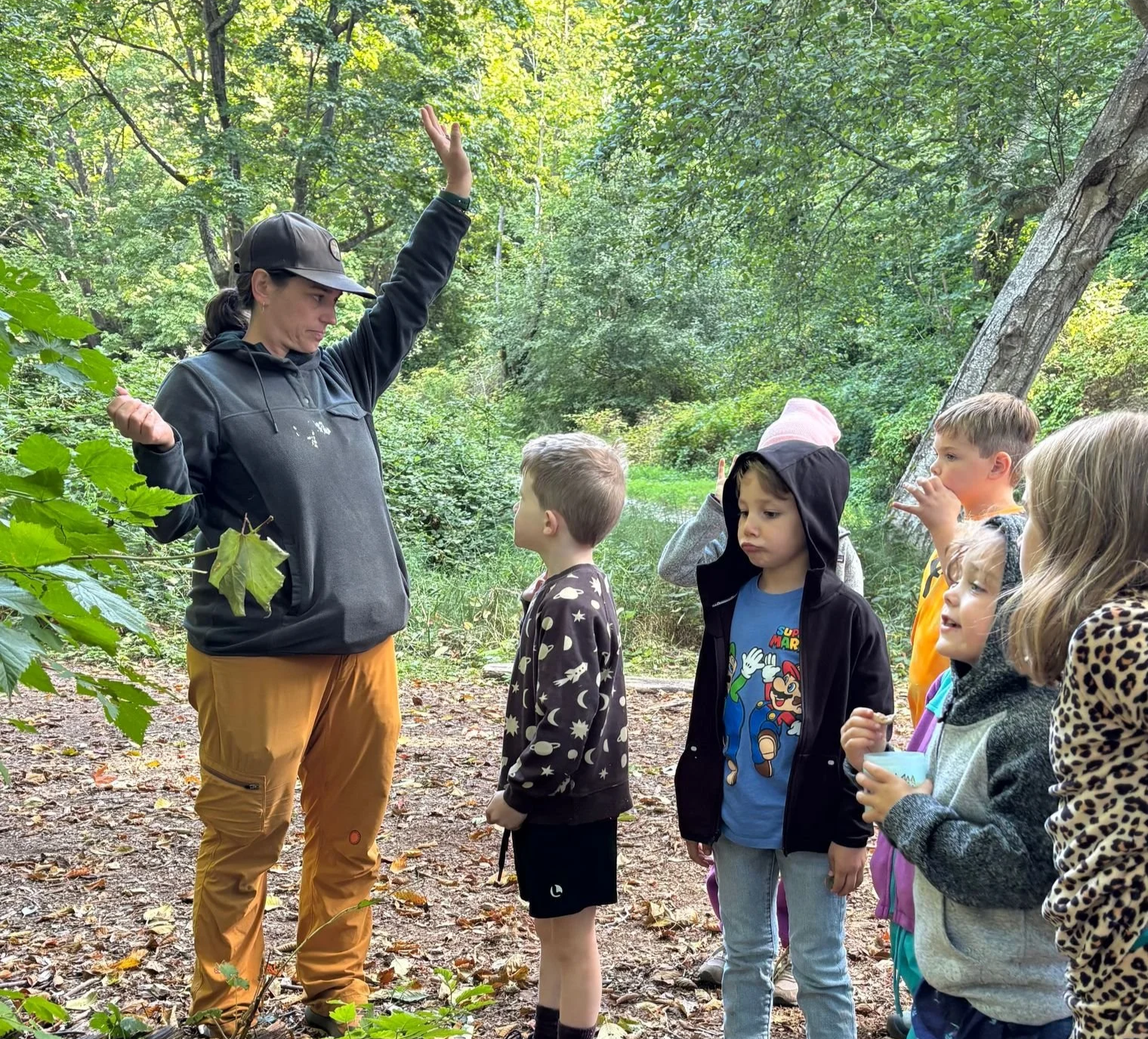 A woman leading a nature educational activity for a group of children in a forest, with trees and green foliage in the background.
