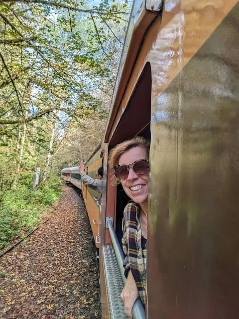 A woman smiling and leaning out of a train window while a person in the background reaches out of another train car, with lush green trees along a railway track.