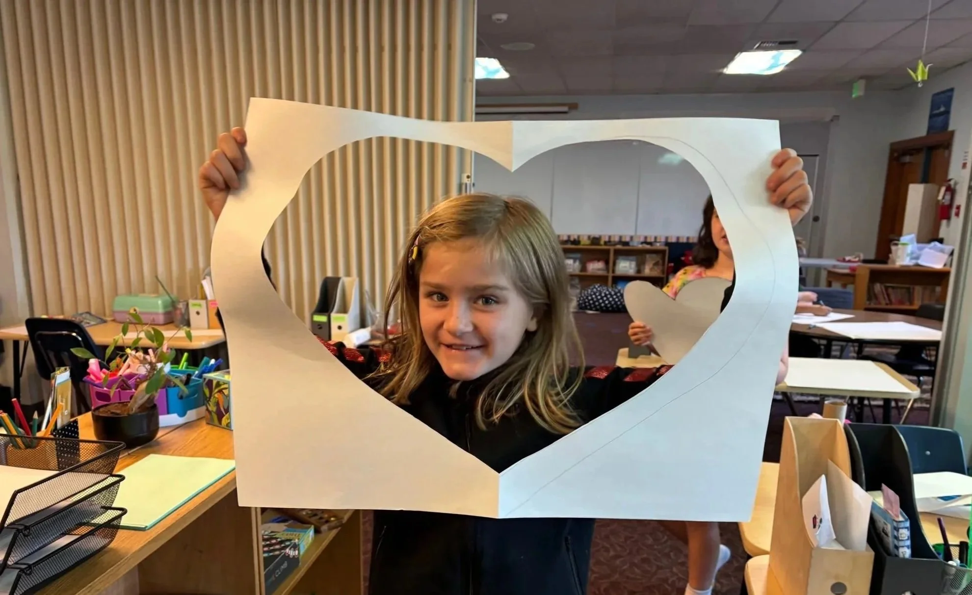 A young girl holding a heart-shaped cutout in front of her face, looking through a large white heart-shaped frame, in a classroom setting.
