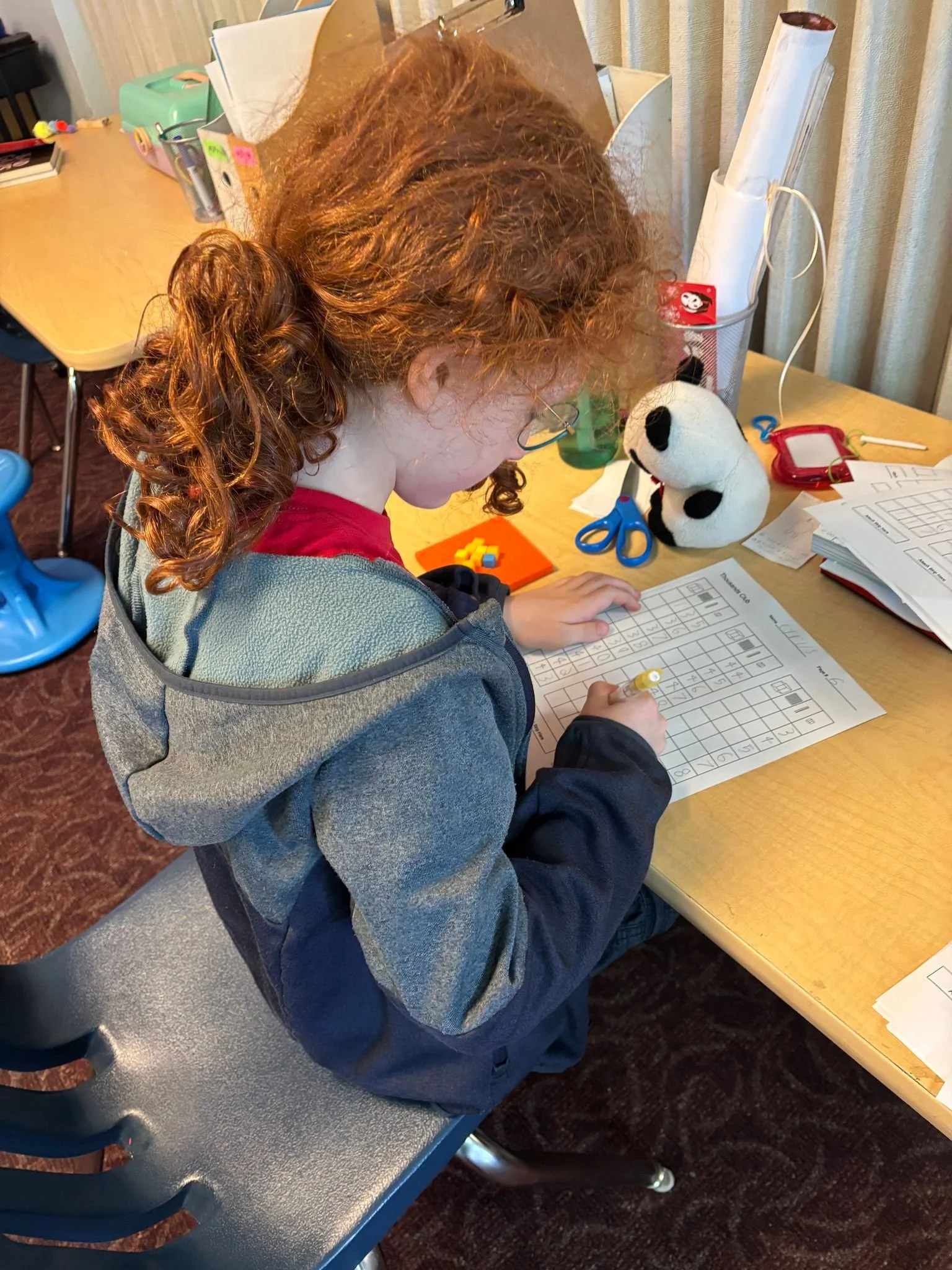 A young girl with curly red hair, wearing a gray and navy jacket, is sitting at a desk doing a worksheet. The desk has various school supplies and a stuffed panda bear.