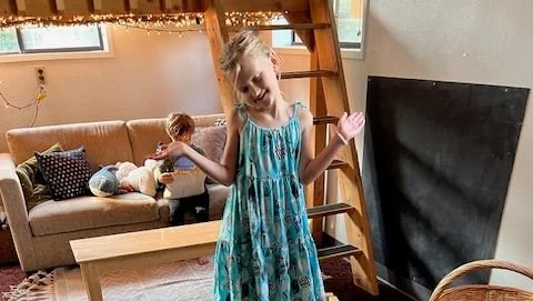 A girl in a blue dress smiling and dancing in a living room with a boy playing with toys on a couch, a ladder, and a chalkboard in the background.