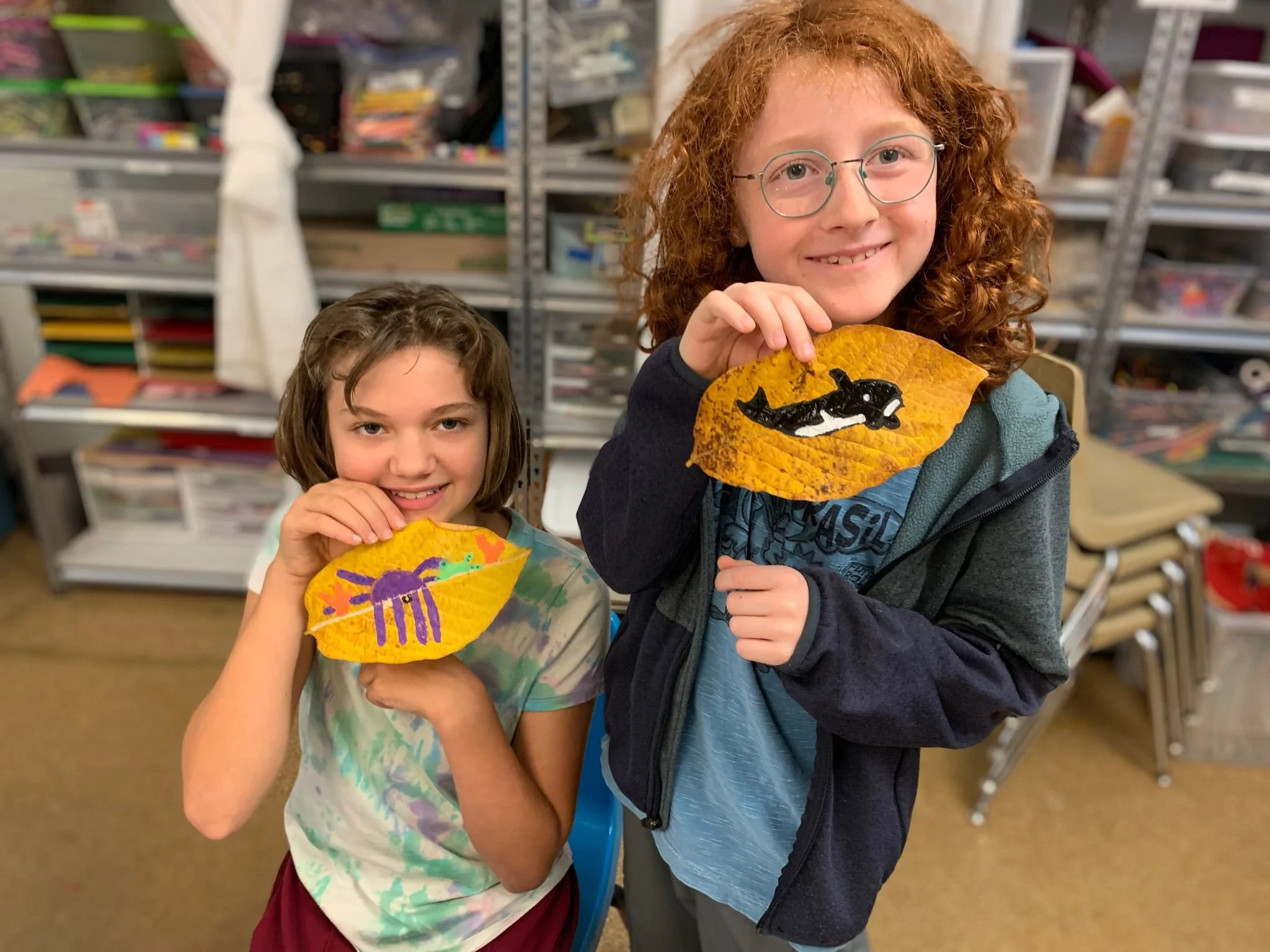 Two young girls displaying leaf art with painted animals. One girl with curly red hair and glasses holds a yellow leaf with a painted orca whale. The other girl with straight brown hair holds a yellow leaf with a painted purple spider. They are in an art classroom with shelves of supplies in the background.