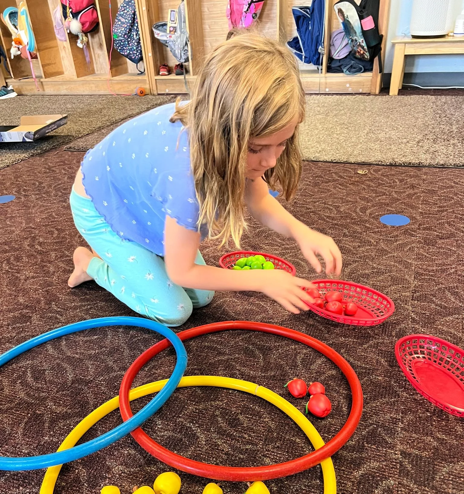 A young girl kneeling on a carpeted floor, organizing small plastic balls into red baskets. She is wearing a light blue shirt and sky blue pajamas with white patterns, with sports hoops and additional balls spread around her.