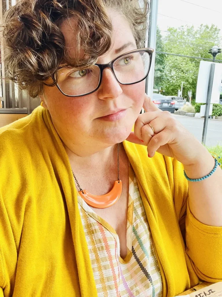 A woman with short curly hair wearing glasses and a yellow cardigan, sitting indoors near a window, with her hand resting on her face, and a necklace with an orange pendant.