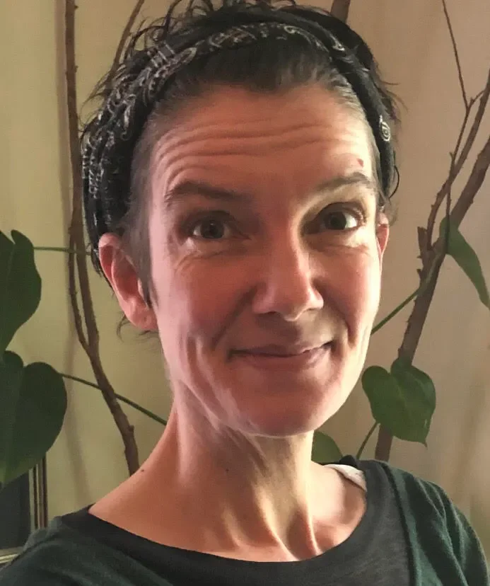 A woman with short dark hair and a black headband smiling in front of leafy houseplants indoors.