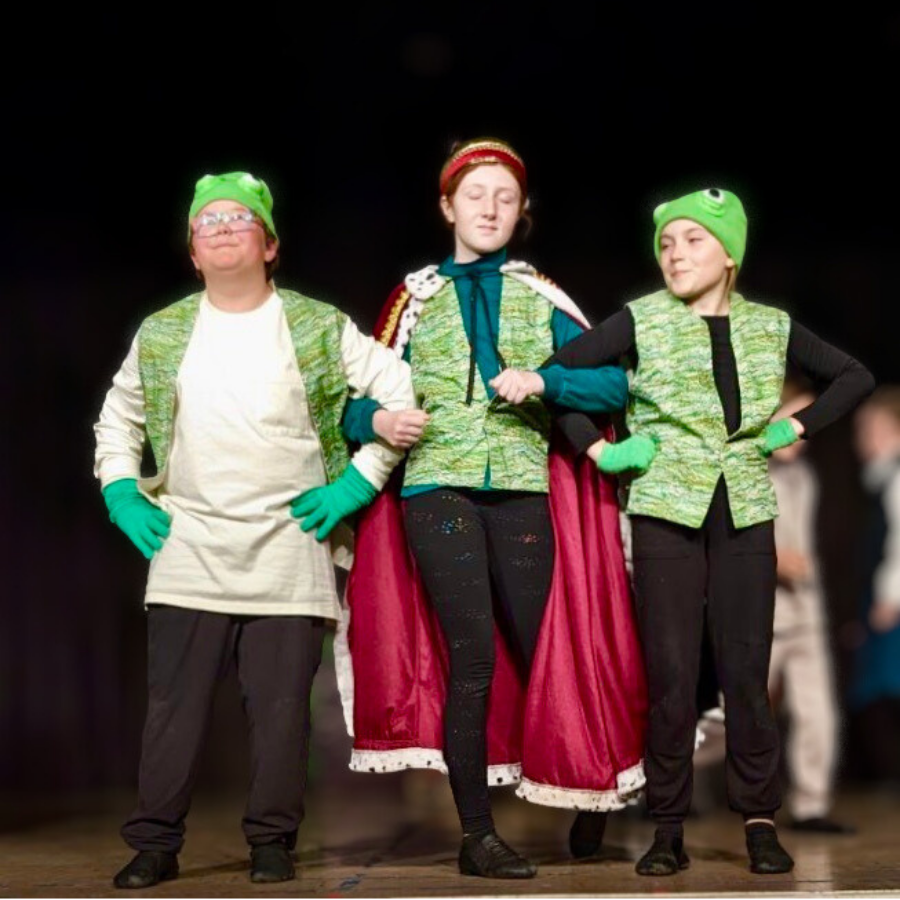 Three children in costumes performing on stage, with two dressed in green frog hats and vests and one dressed as a princess or queen with a red cape and crown.