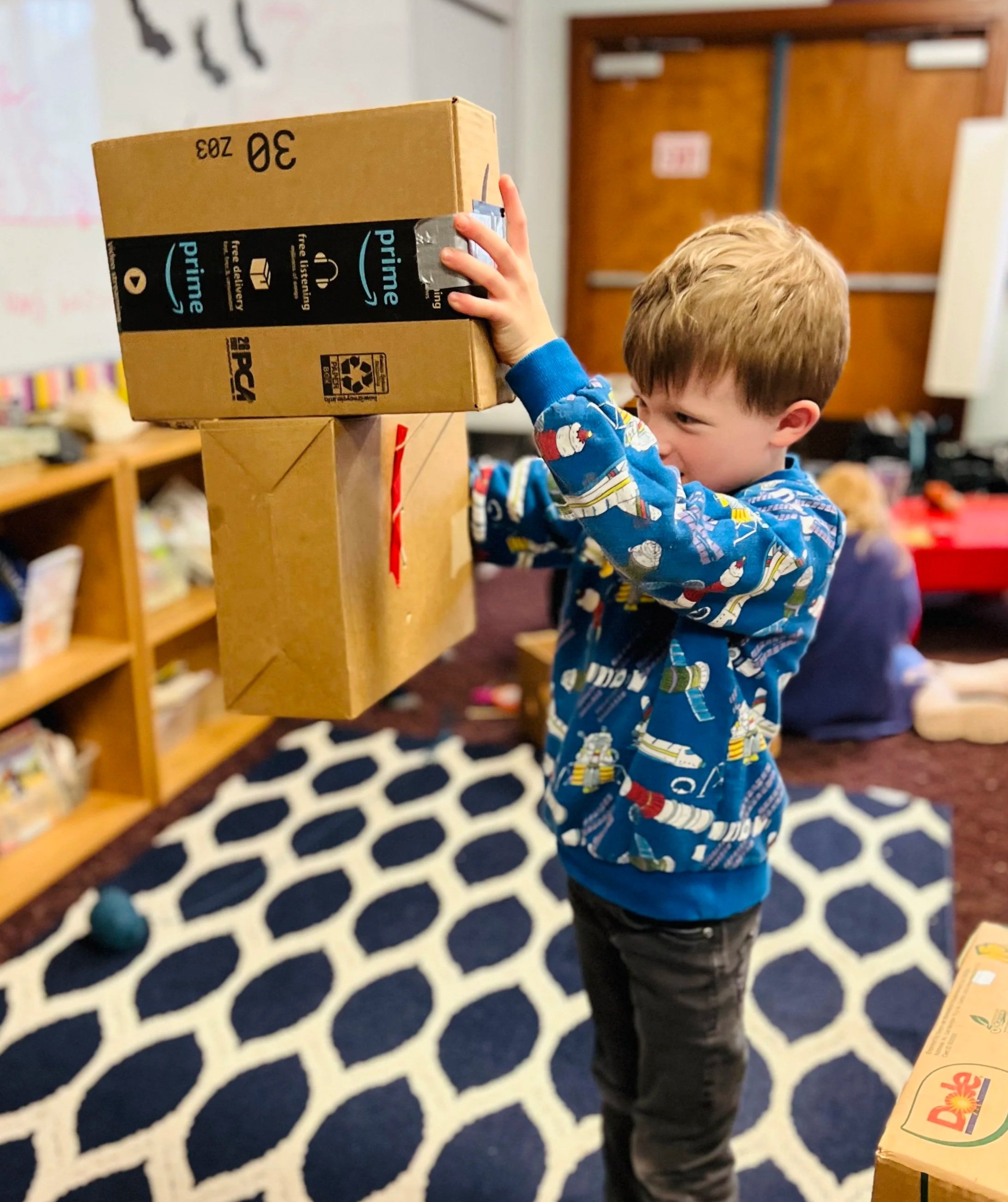 A young boy wearing a blue sci-fi themed pajama top and black pants is holding a cardboard box with Amazon Prime branding over another box. The background shows shelves with various items and a patterned rug on the floor.