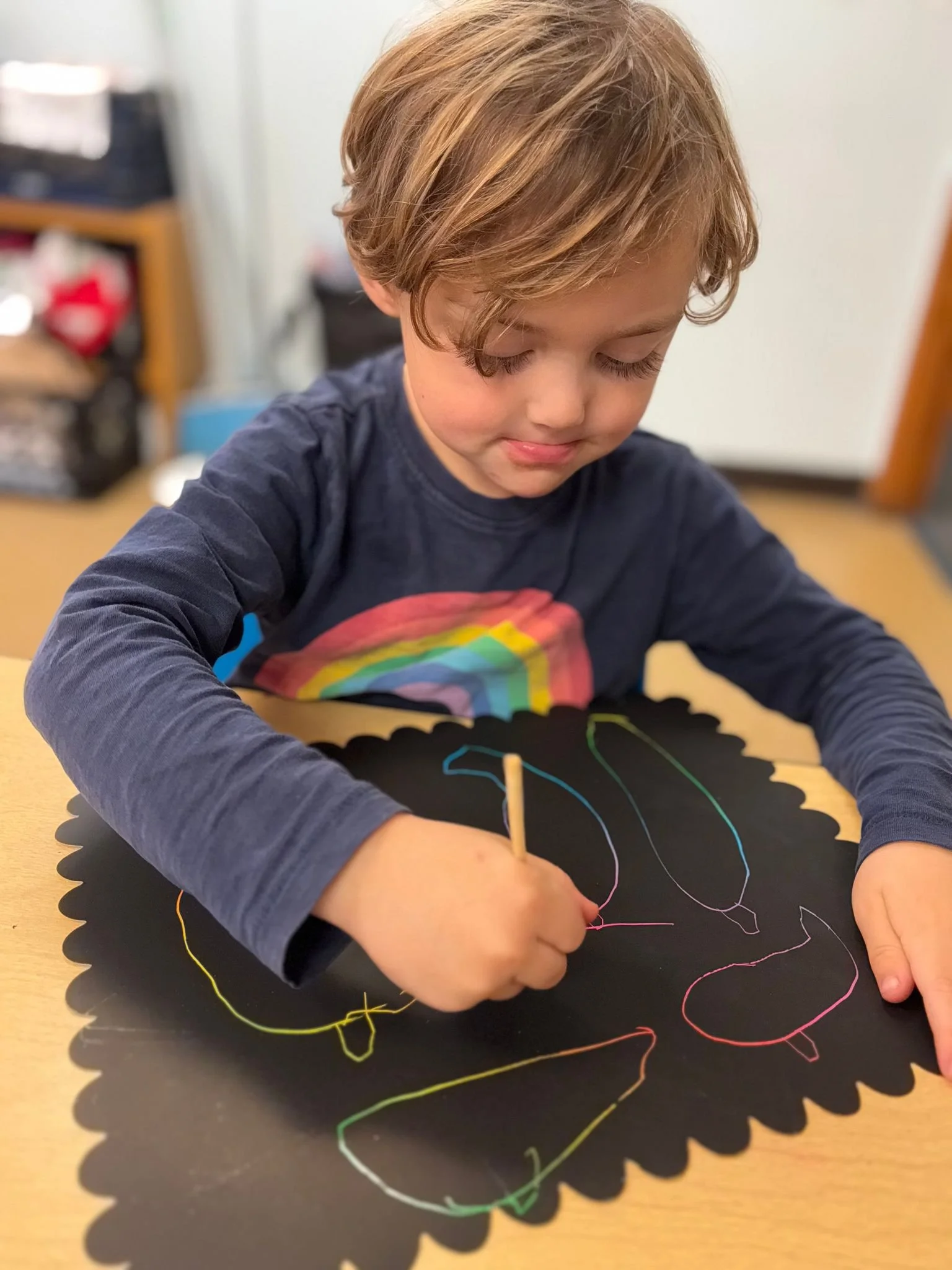 A young child with light brown hair in a dark long-sleeve shirt is drawing colorful squiggly lines with a chalk on black paper.