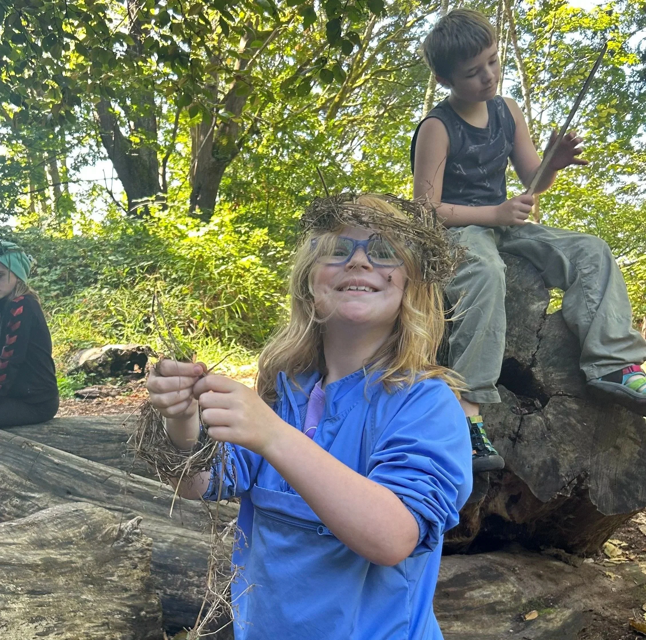 Three children in a forest; one girl with glasses and a blue jacket holding a dirt nest, smiling at the camera, another boy sitting on a large tree stump in a sleeveless shirt, and a third child partially visible sitting on a log.