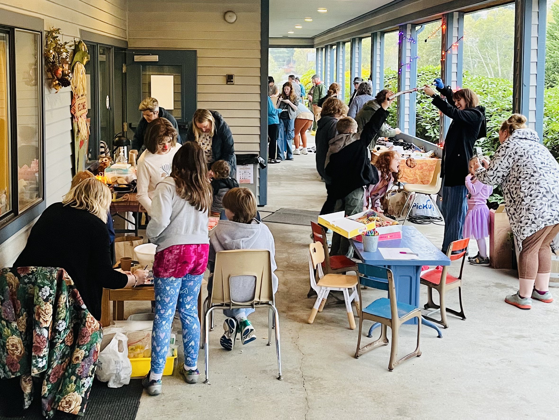 Children and adults at a festive outdoor craft and activity market on a porch, with tables displaying crafts and games, some people line up at a table, and the scene is decorated with colorful lights.