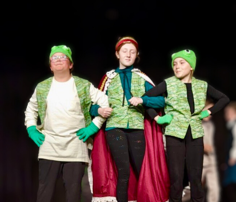 Three children are on stage, dressed in costumes resembling characters from the movie 'Inside Out,' including green hats and gloves, with one wearing a cape, standing in front of a dark background.