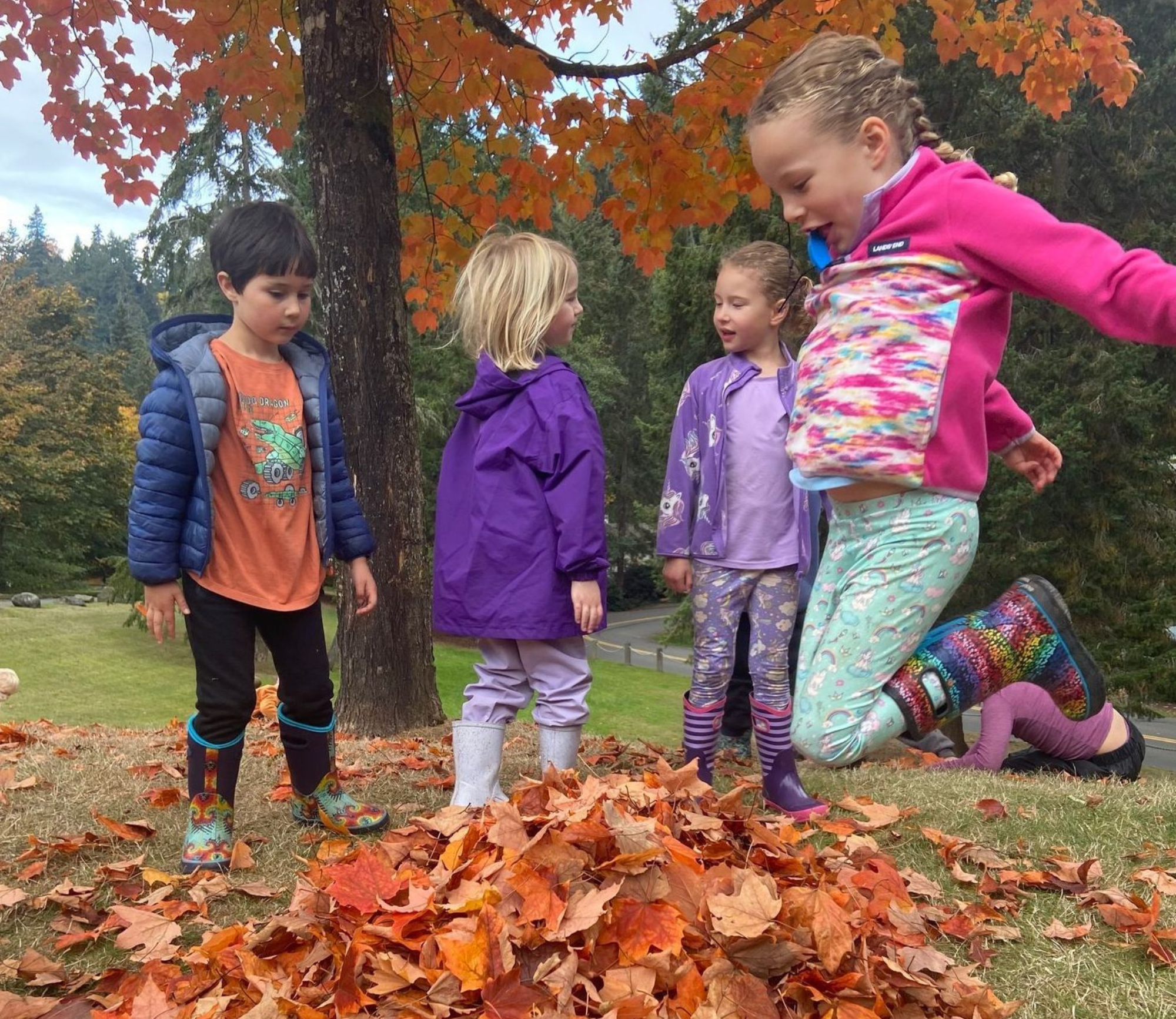 Four children and one adult in colorful rain gear playing in a pile of fallen autumn leaves outdoors during daytime.