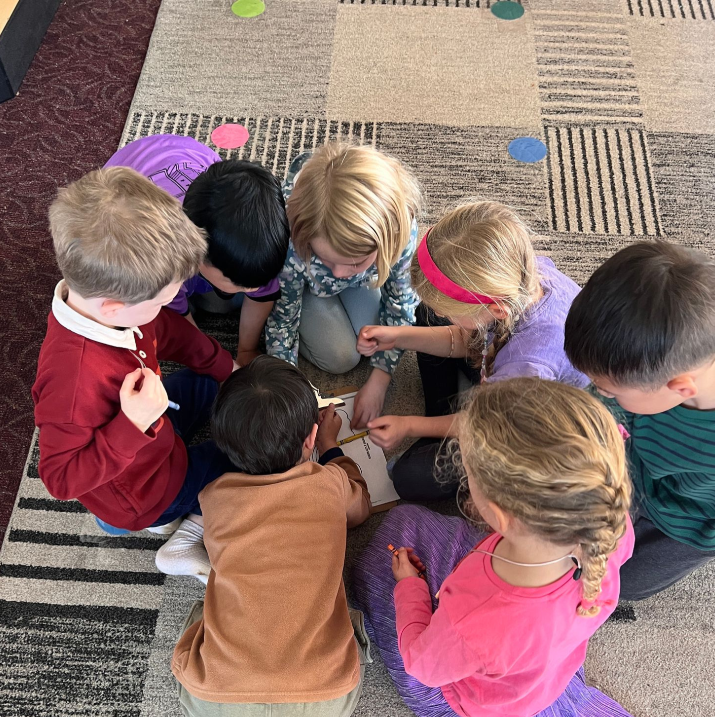 A group of children sitting on a carpeted floor, gathered around a large sheet of paper, engaging in a collaborative activity or game.