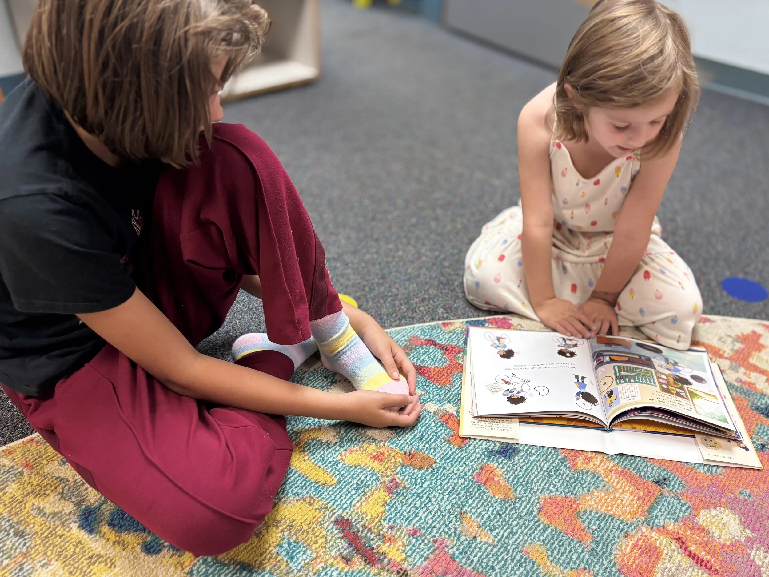 Two children and an adult sitting on a colorful rug, reading a book together on the floor in a daycare or classroom setting.