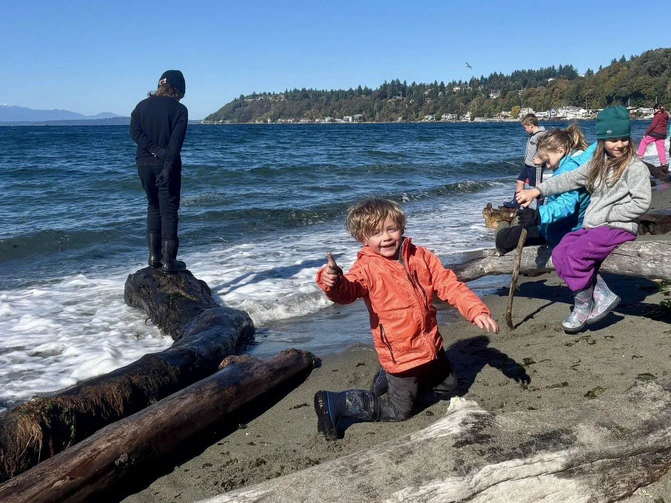 Children playing on the beach near logs with ocean and hills in the background, one child kneeling in the sand giving a thumbs up, others sitting on logs, some fishing or exploring.