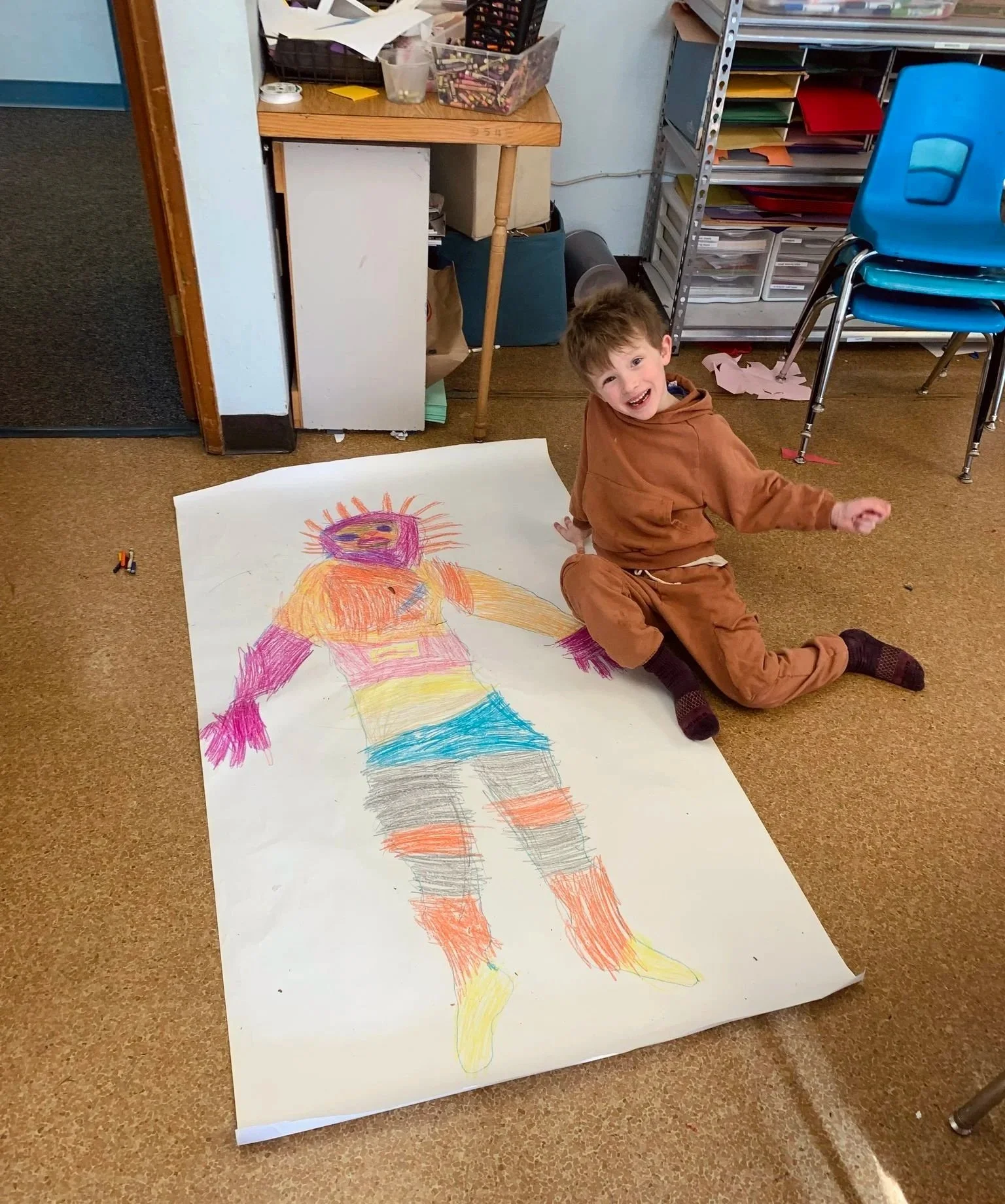 A young boy is sitting on the floor next to a large colorful drawing of a person on a large sheet of paper. He is smiling and wearing a brown hoodie and brown pants. The background shows shelves with colored papers and chairs.