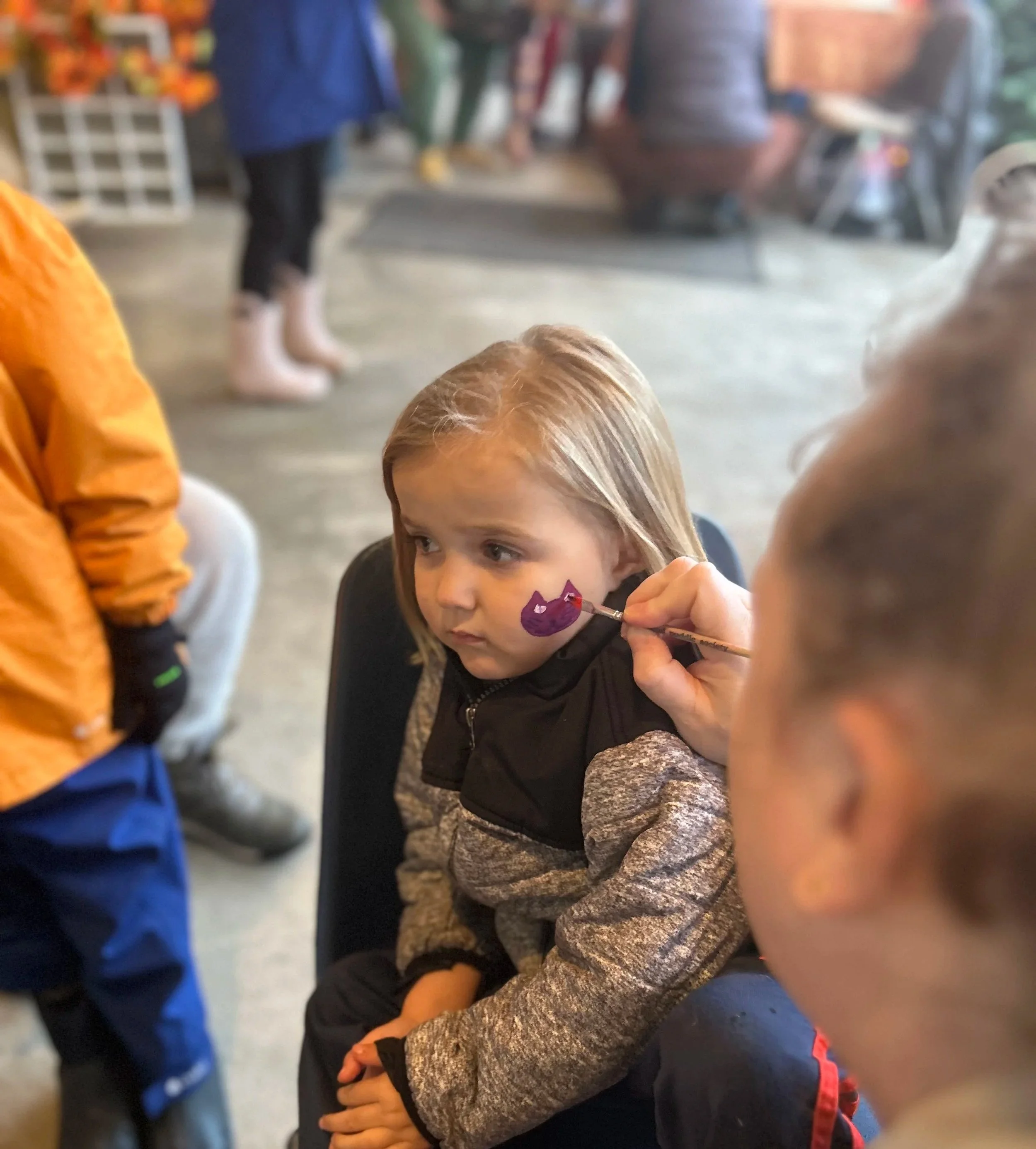A young girl sitting in a chair while an artist paints a purple face on her cheek. The girl has blonde hair and looks slightly to the side with a serious expression. Others are visible around her, and the background shows a busy indoor setting.