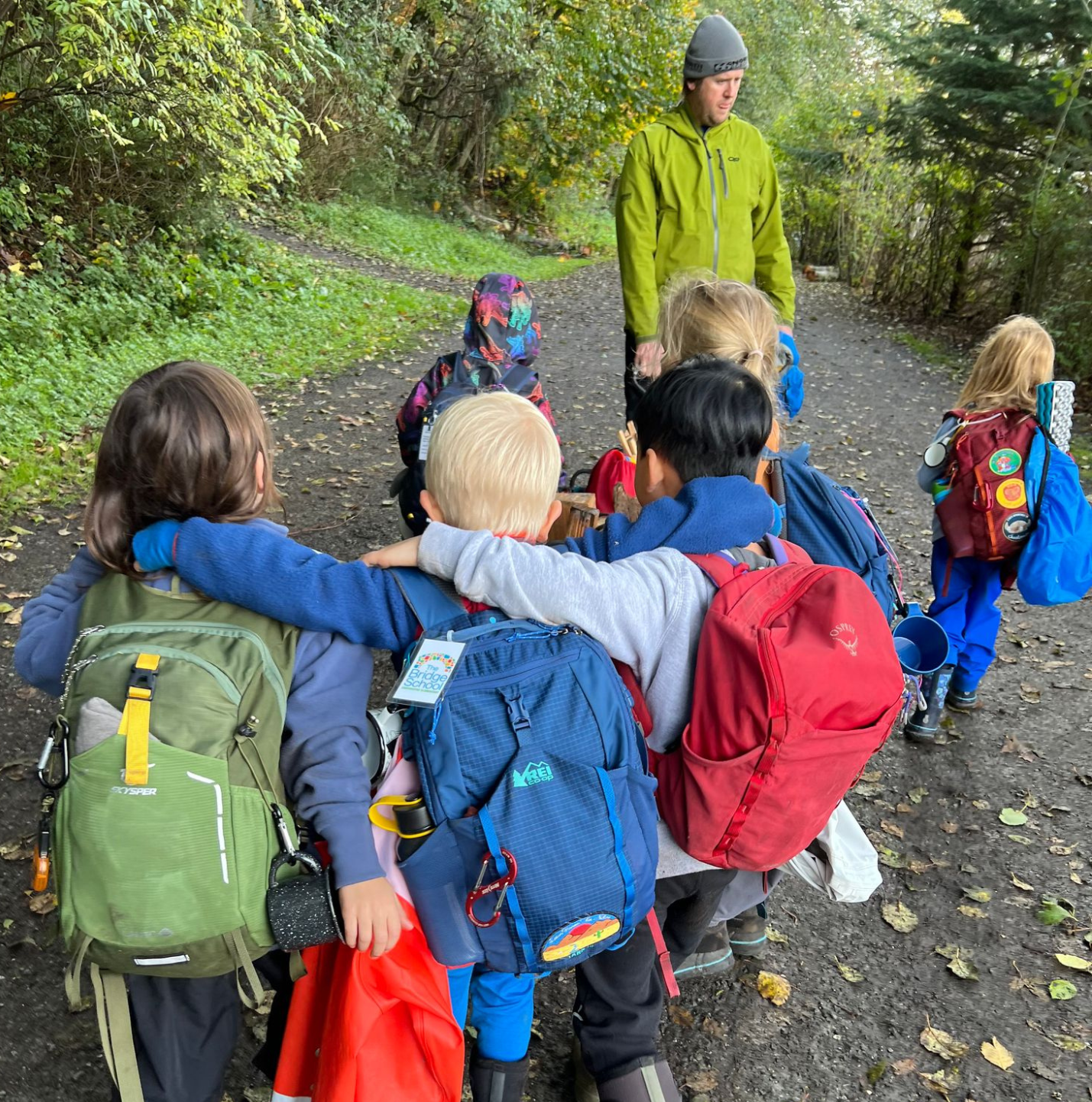 A group of children with backpacks on a forest trail, with an adult supervising.