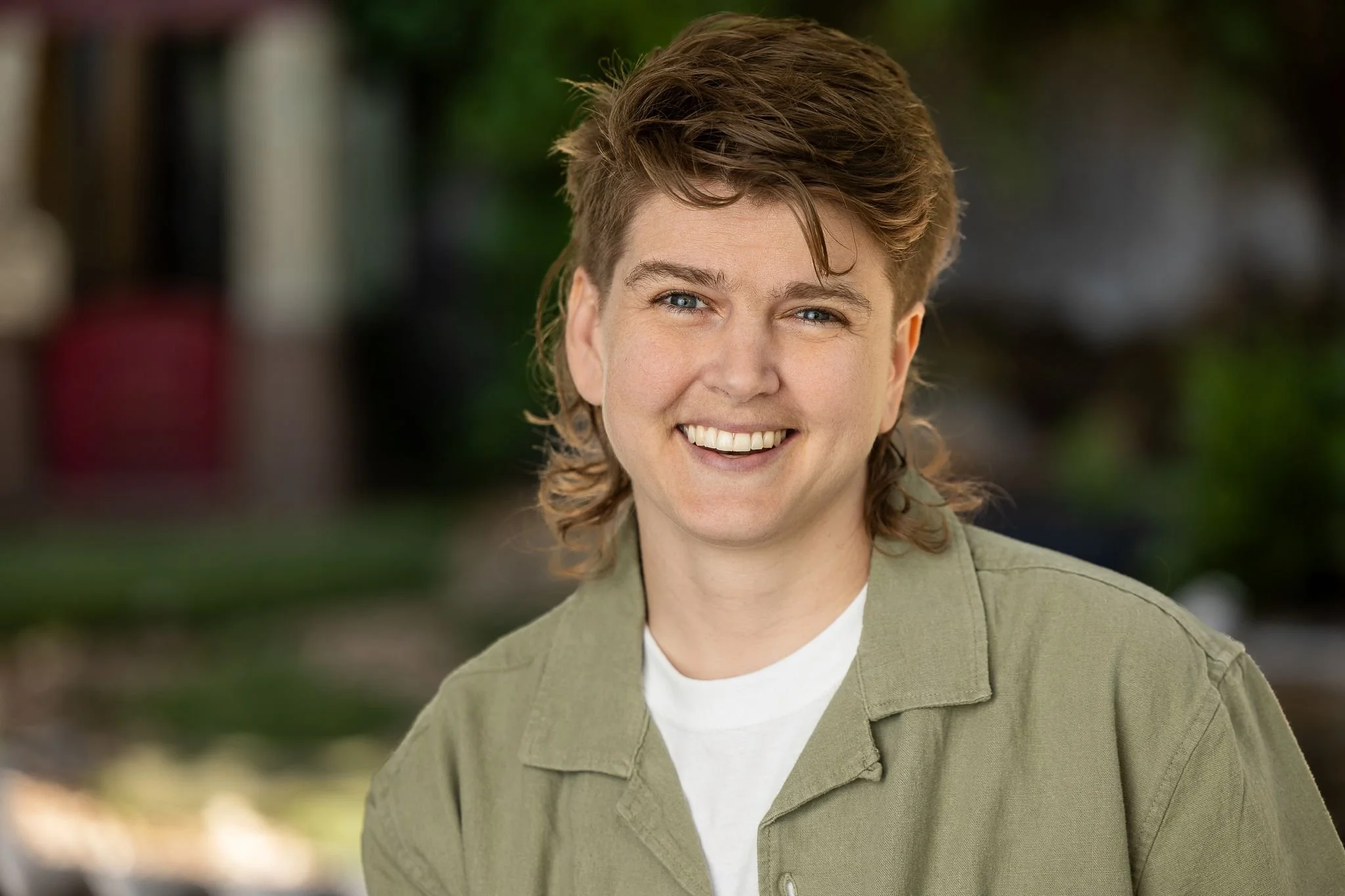 A smiling woman with short, wavy brown hair, wearing a light green jacket and white shirt, outdoors with blurred greenery in the background.