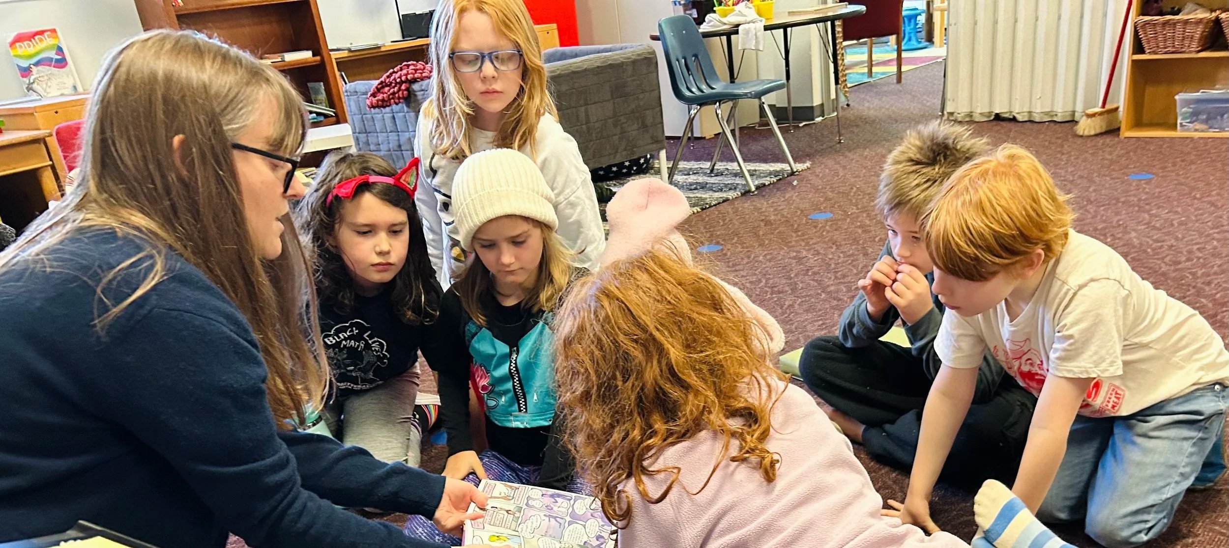A group of children and a woman sitting on the floor in a room, gathered around a comic book or magazine. The woman is showing the book to the children, who are attentively looking at it. The children are diverse in appearance, with some wearing glasses, hats, or costumes, and the room has shelves, chairs, and toys in the background.