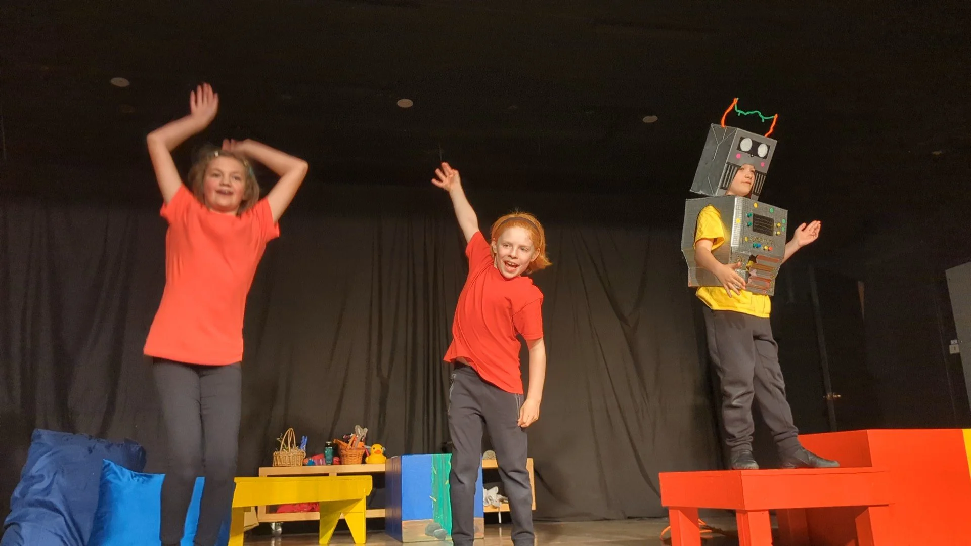 Three children on stage, two girls performing dance or play, one boy dressed as a robot with boxes and wires on stage