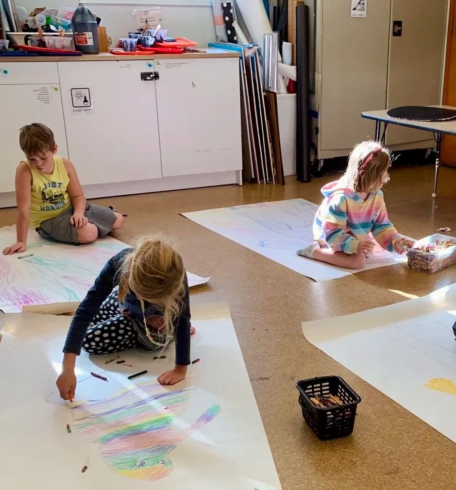 Three children are coloring on large sheets of paper spread on the floor in a classroom, with art supplies and storage cabinets visible in the background.