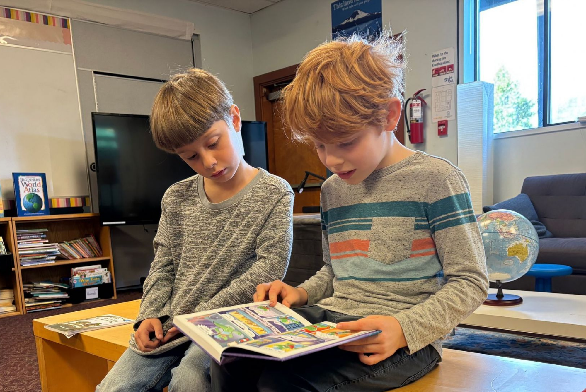 Two young boys sitting on a wooden table in a classroom or library, looking at a colorful book or comic book together.