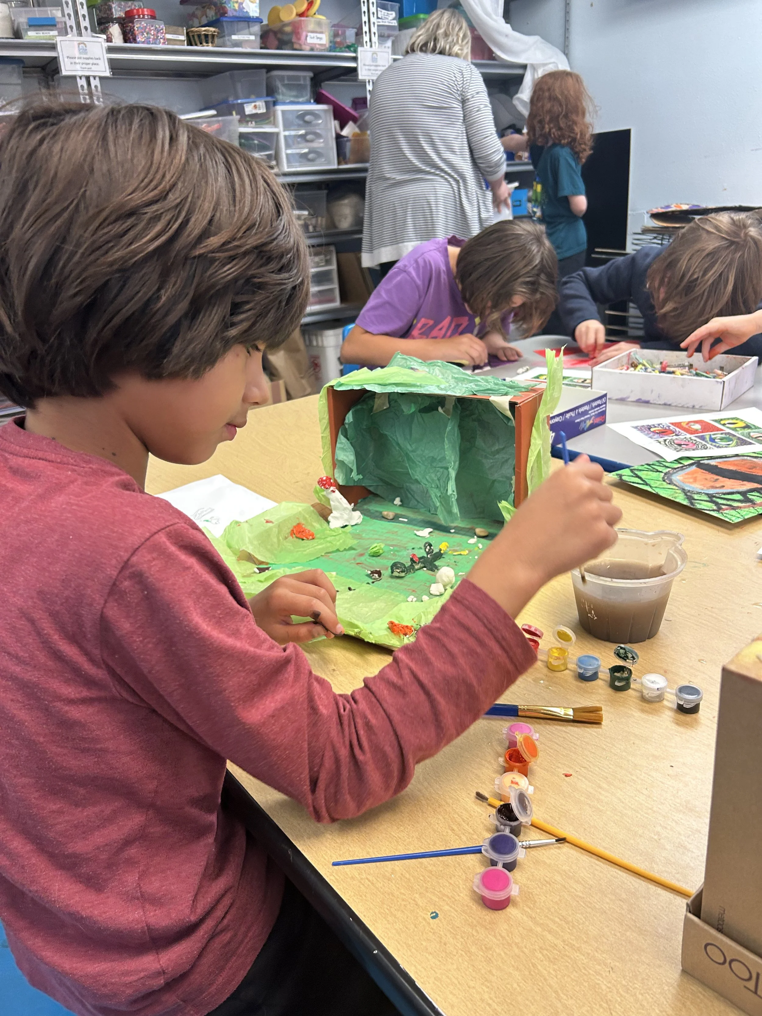 Children painting and crafting at a table in an art classroom with supplies and shelves in the background.