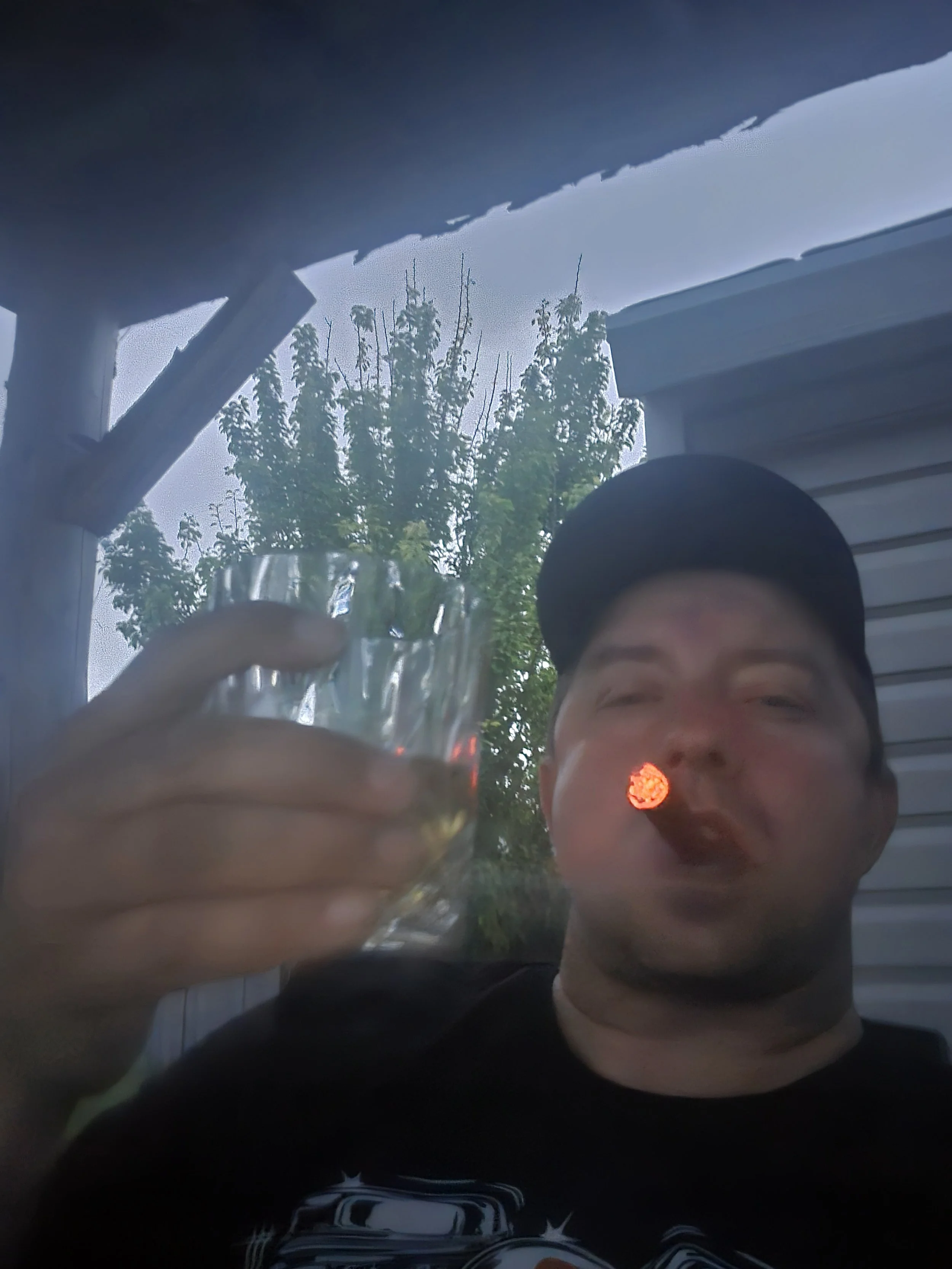 A man wearing a black cap holding a glass of beverage, sitting outdoors with trees and a house in the background, as he smokes a cigarette.
