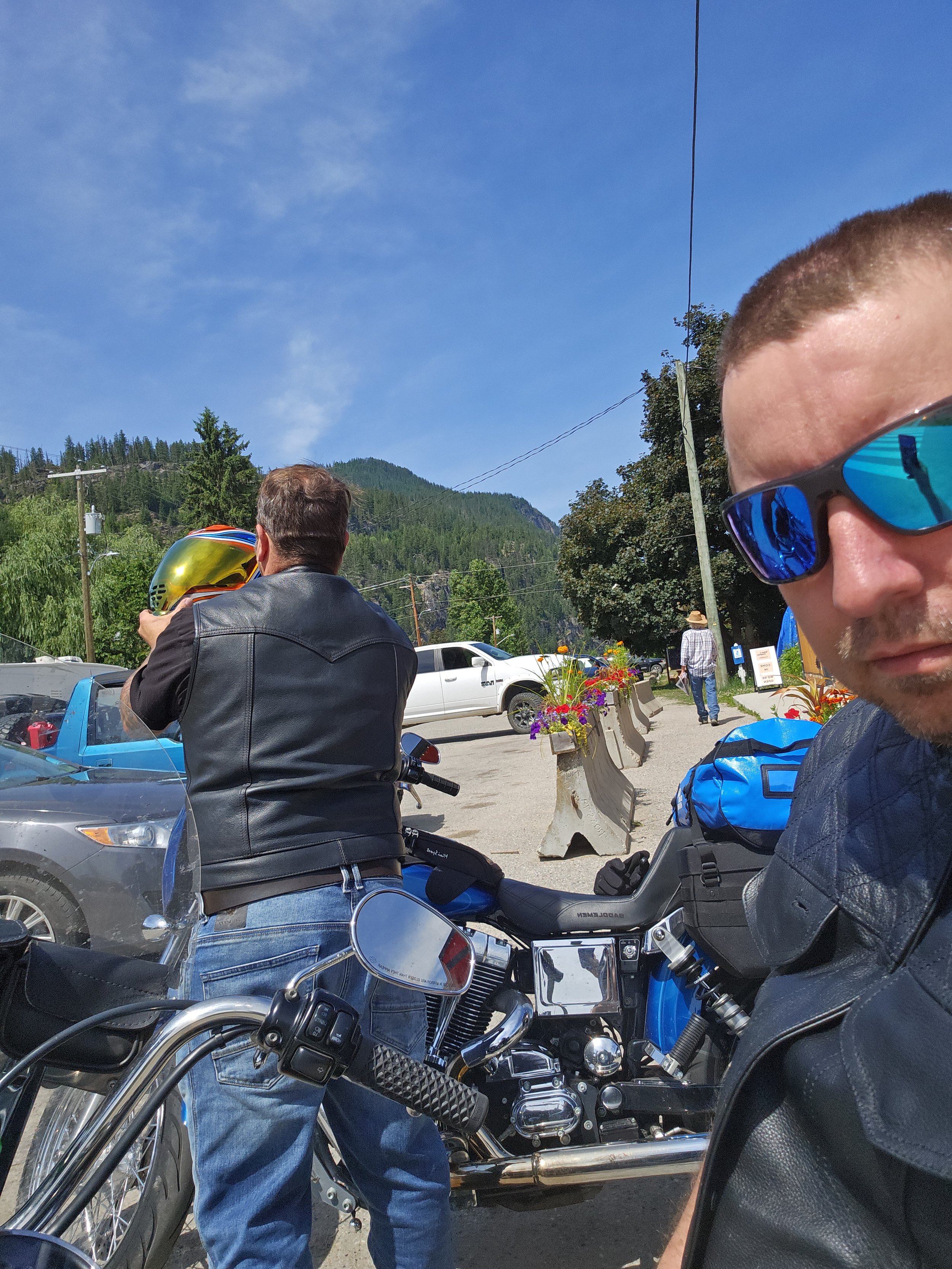 A man wearing sunglasses and a leather jacket taking a selfie, with another man adjusting a motorcycle helmet, parked cars, and mountains in the background on a sunny day.